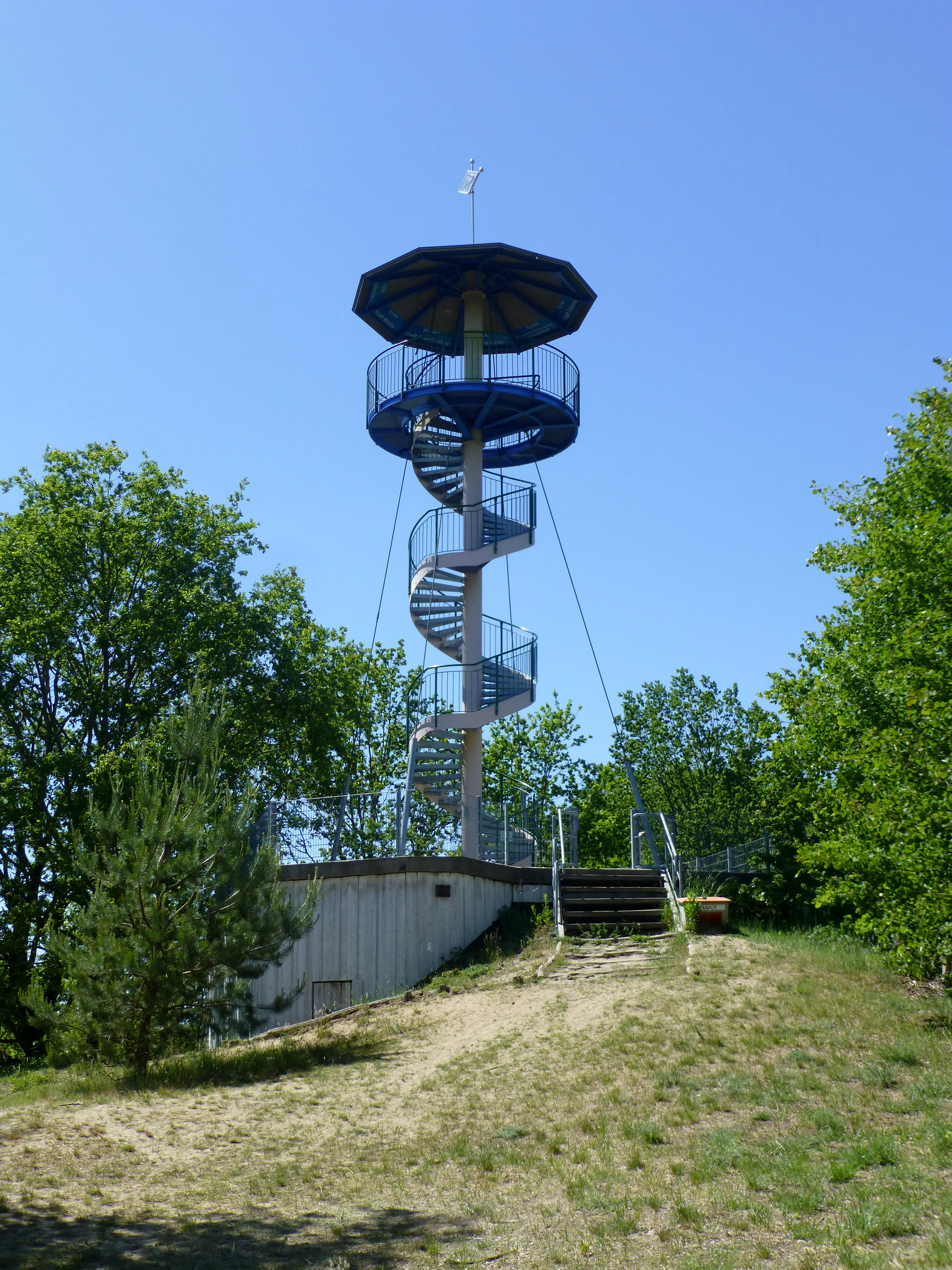 Una torre de observación moderna de espiral de acero se alza sobre una pequeña colina arenosa rodeada de árboles caducifolios verdes bajo un cielo azul despejado.