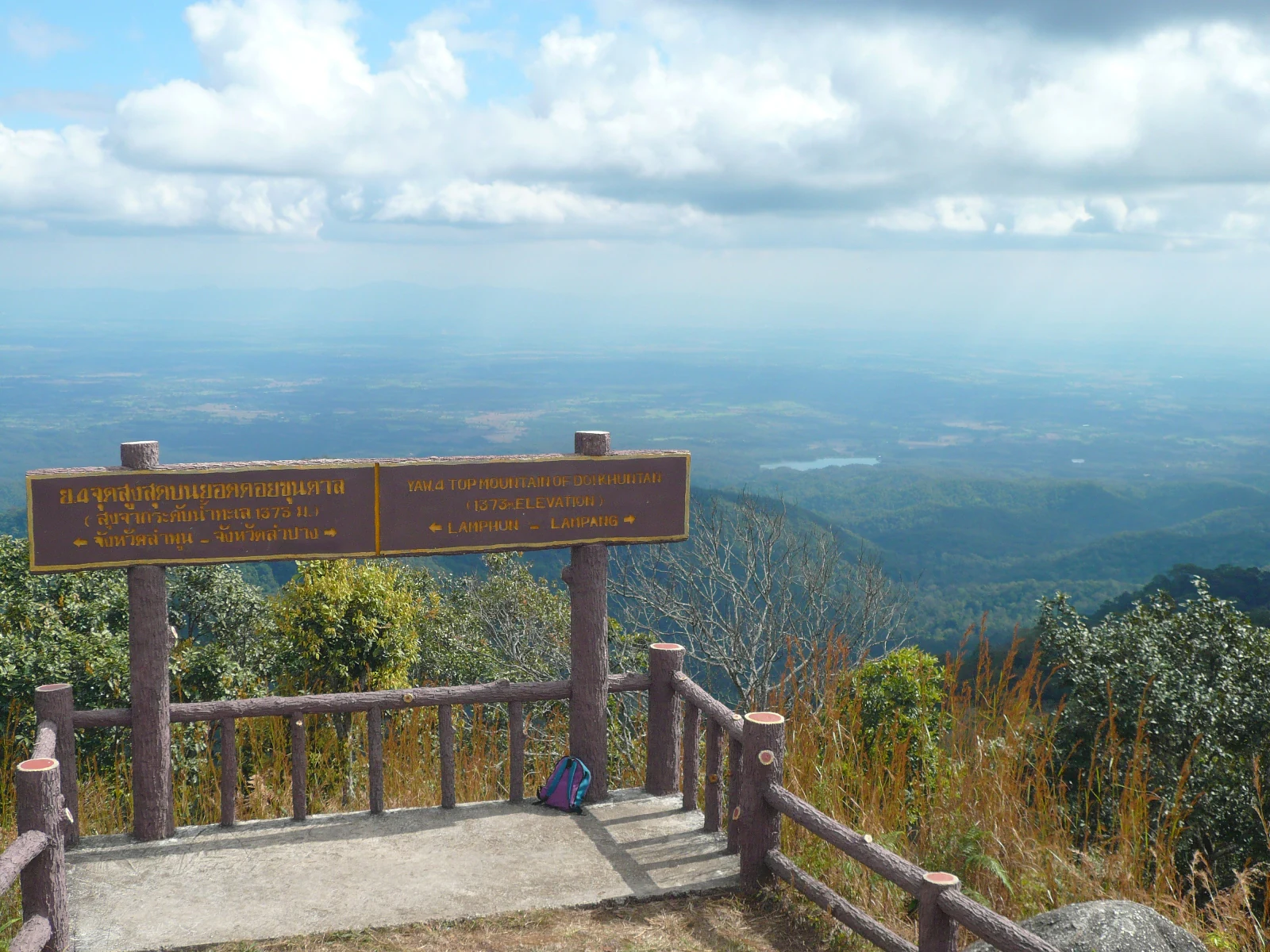 Wooden sign with Thai text at a mountain observation deck overlooking valley and distant mountains