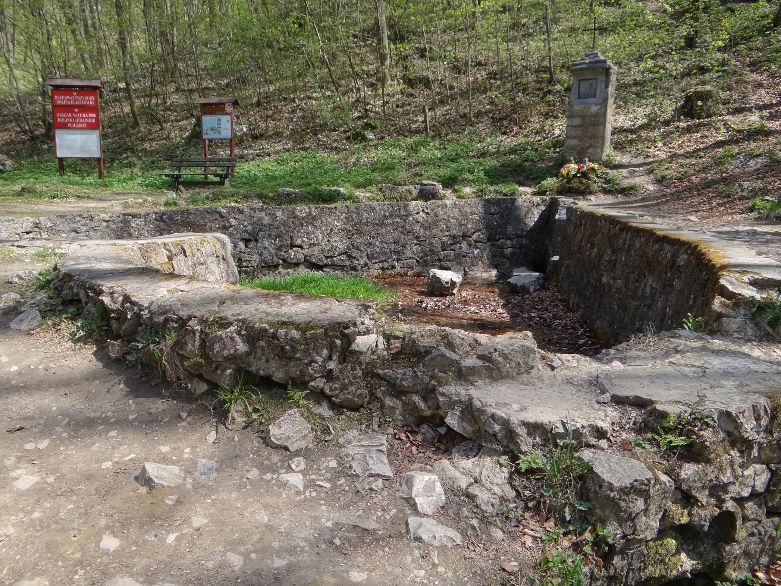 A stone-walled spring enclosure set into a hillside in a forest, featuring a small stone shrine to the side and information signs in the background.
