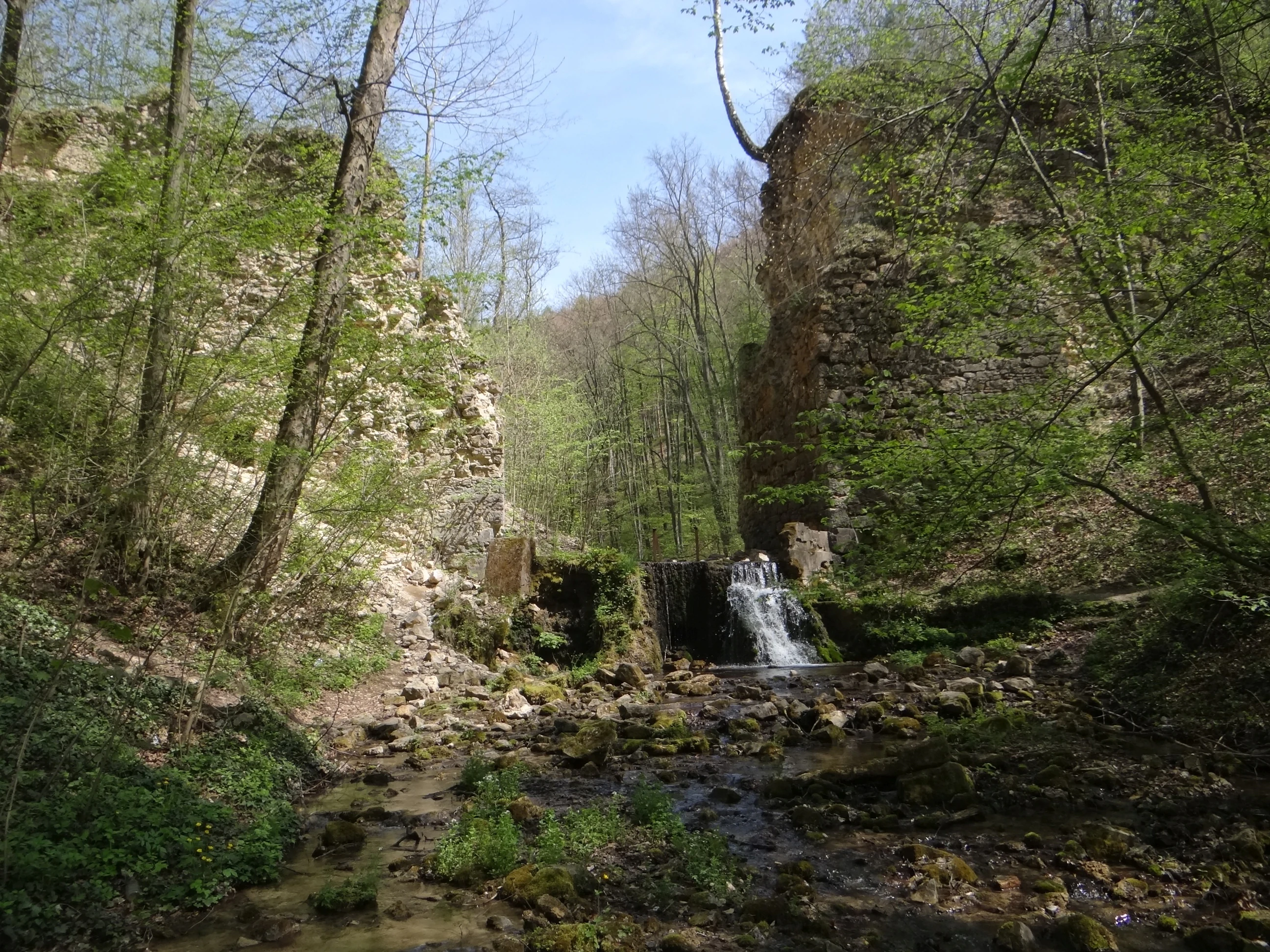 Ancient stone arch ruins situated over a small stream in a dense forest, with a small waterfall flowing between the stone pillars.