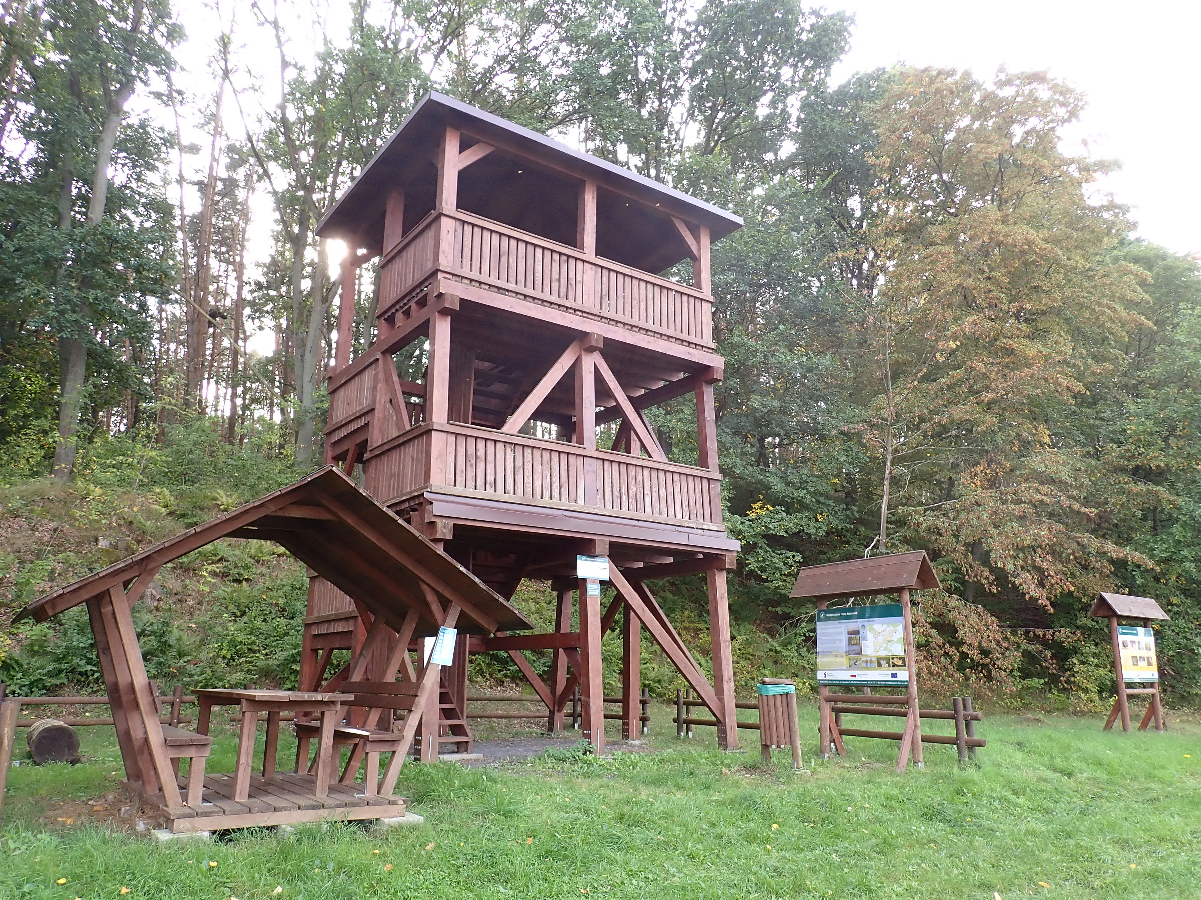 A multi-level wooden observation tower stands at the edge of a forest, next to a small wooden picnic structure and interpretive signage.