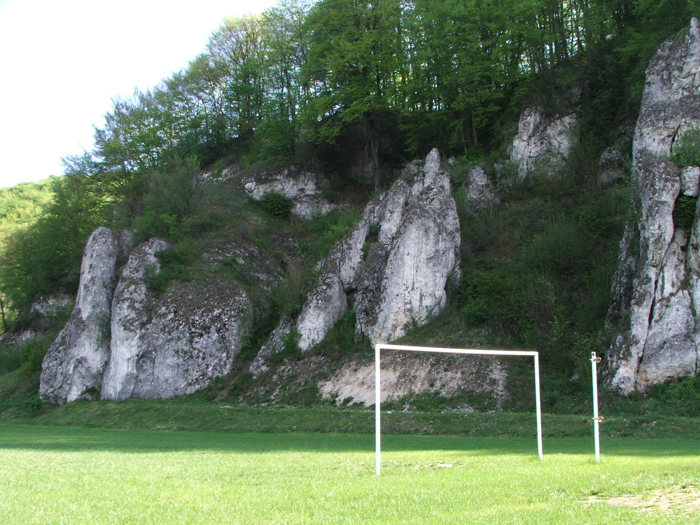 A view of steep white limestone cliff formations rising above a green grassy field with a white soccer goal frame in the foreground.