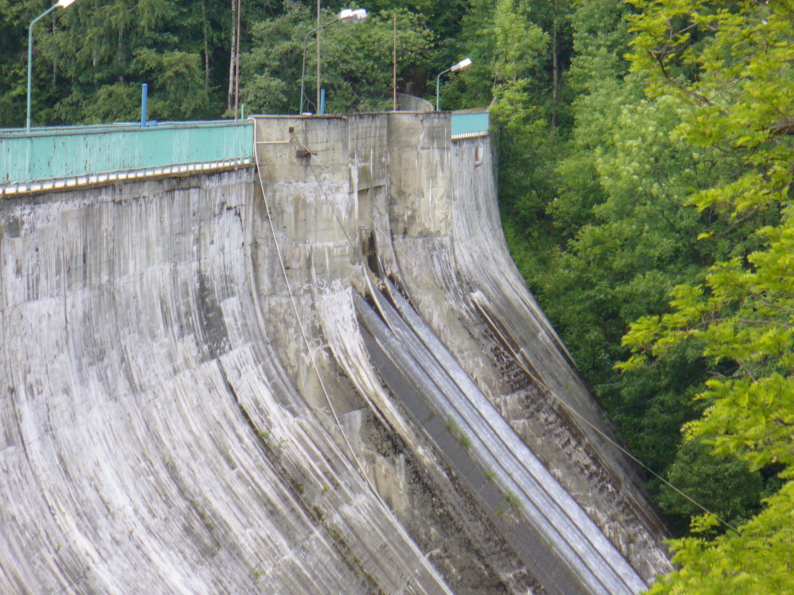 A close up view of the curved, weathered concrete wall of the Wapienica Dam, surrounded by lush green trees.