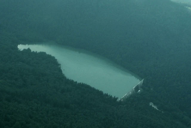 An aerial photograph shows a water reservoir nestled within a dense forest, with a prominent dam structure visible at one end.