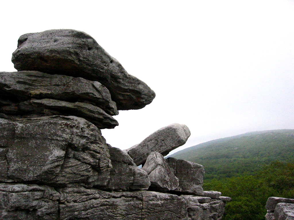 A close up view of a stacked, weathered gray sandstone rock formation in the foreground, with a hazy, green forested mountain slope in the background under an overcast sky.