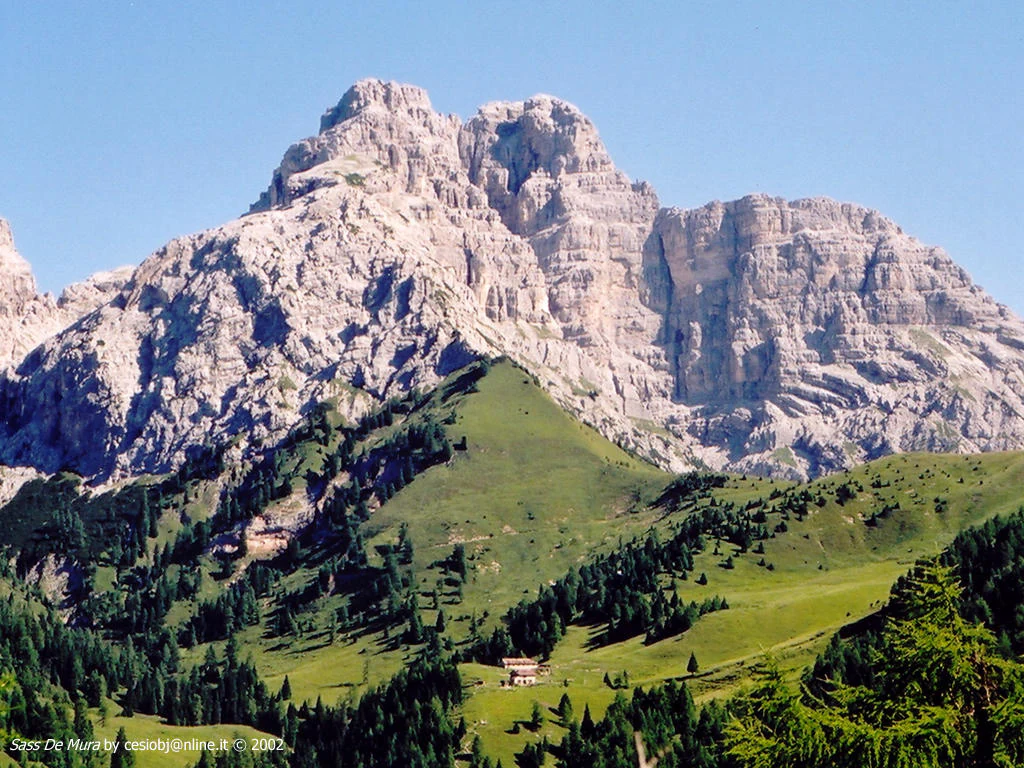 Rocky mountain peak with green slopes and forests below, featuring a small building in a meadow