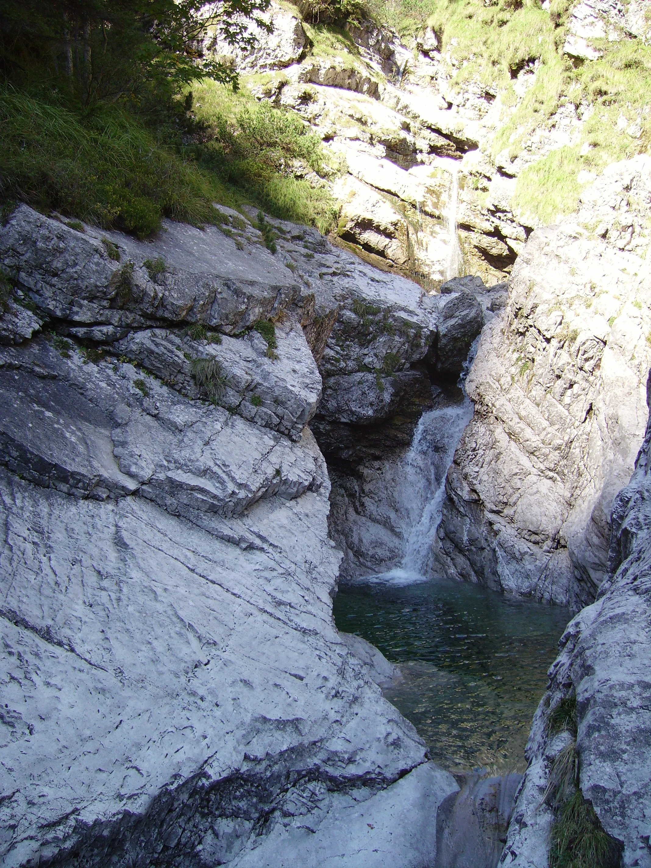 A small waterfall cascading into a rocky pool surrounded by green vegetation and stone cliffs