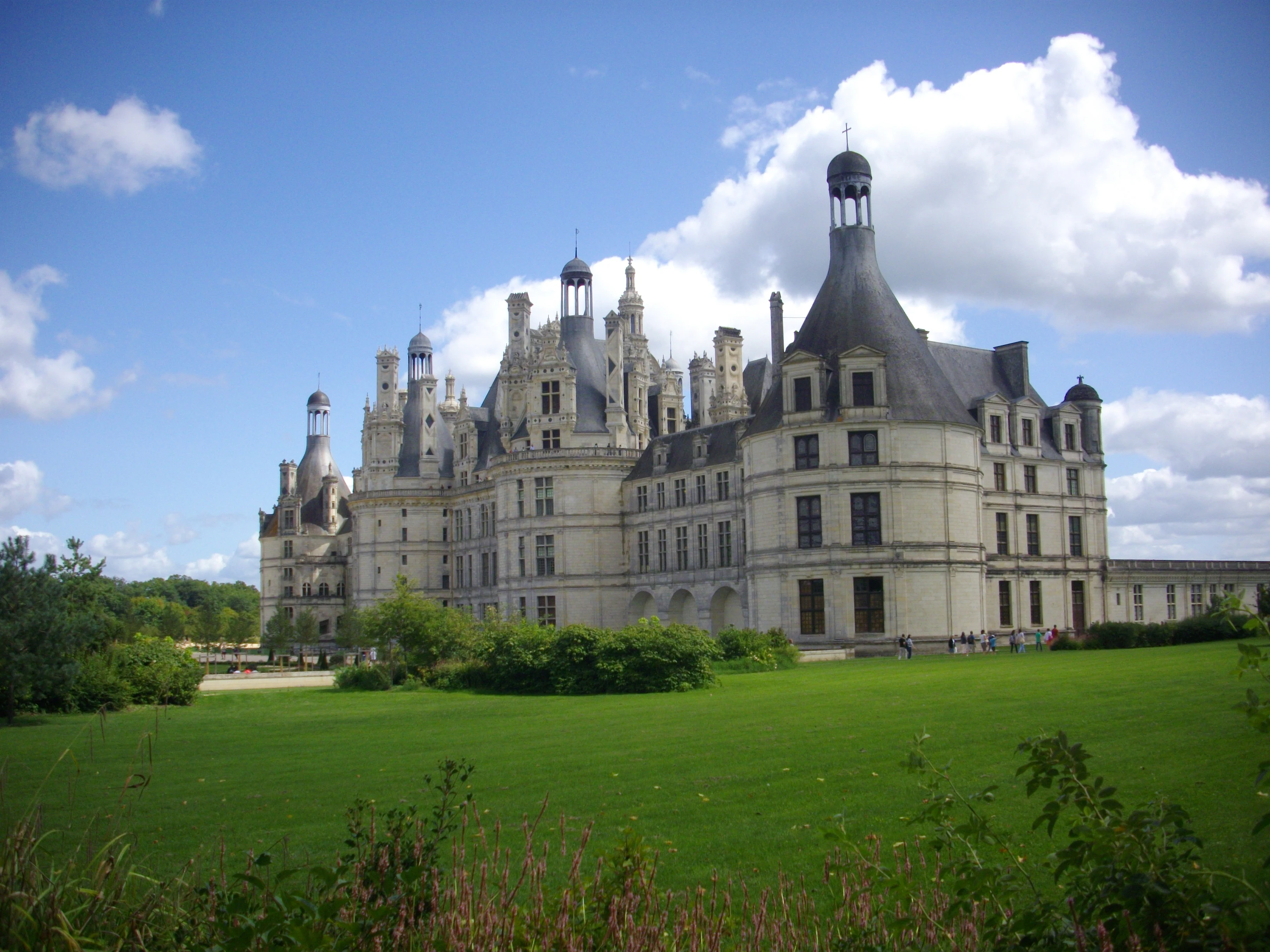 A wide view of the grand Renaissance style Chateau de Chambord in France, featuring white stone towers, multiple roof spires, and dark grey roofing, surrounded by lush green lawns under a blue sky with white clouds.