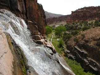 A waterfall flows down a rocky slope with red canyon walls and green vegetation visible in the background