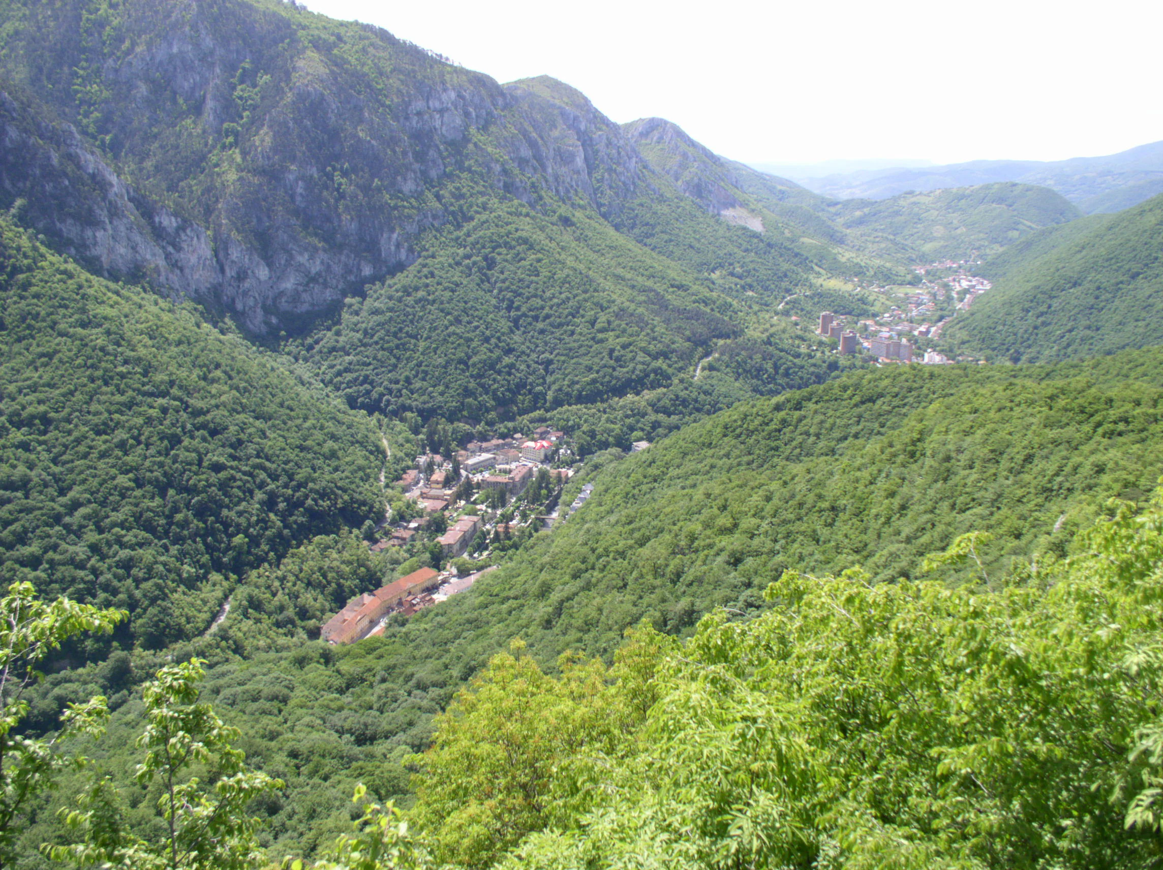 Panoramic view of Baile Herculane town nestled in a lush green valley surrounded by mountain ranges with dense forests and clear sky