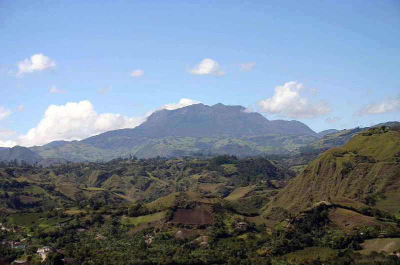 Wide landscape view showing rolling green hills in the foreground and Doña Juana Volcano in the background under a clear blue sky with scattered clouds