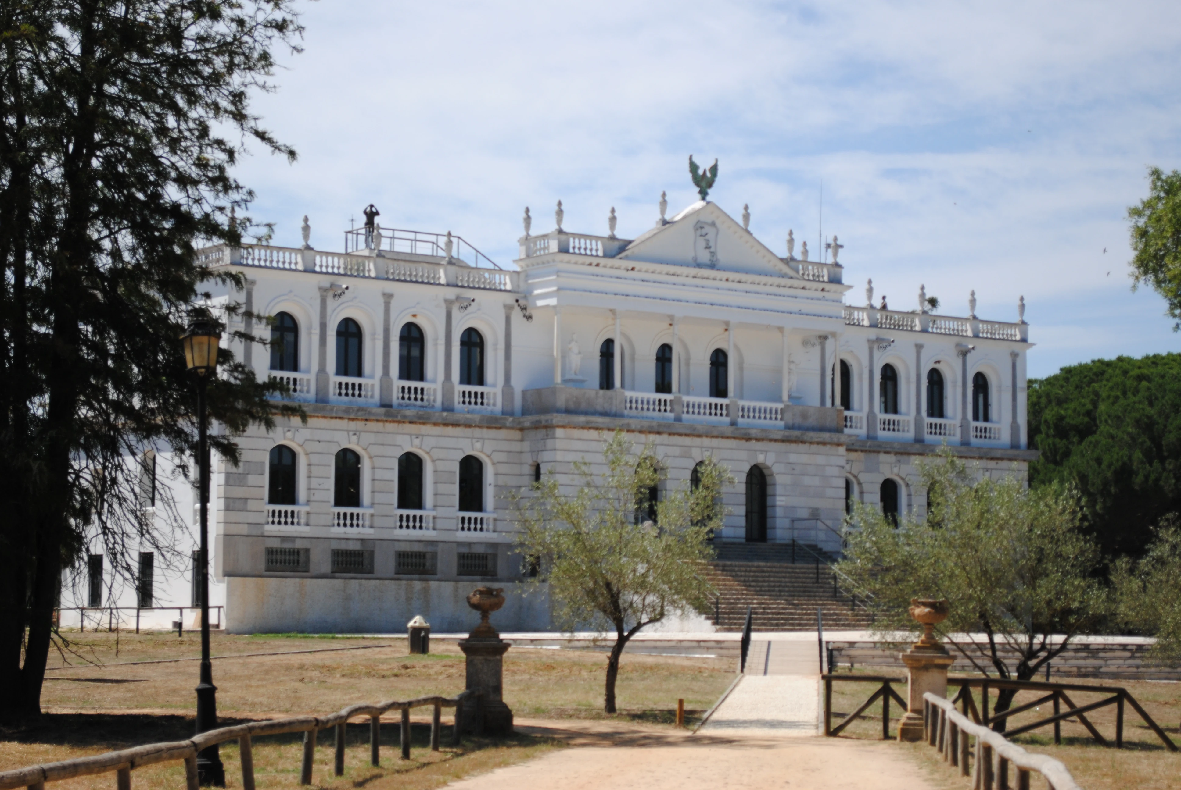 White historic building with multiple windows and balconies, an eagle statue on top, surrounded by trees, grass, and a pathway with wooden fences