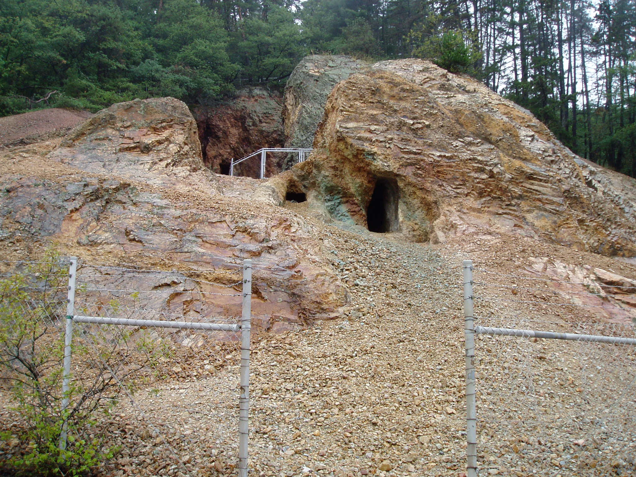 A view of the entrance to an old, small copper mine carved into a rocky, reddish-brown rhyolite hillside, secured with a chain-link fence in the foreground.