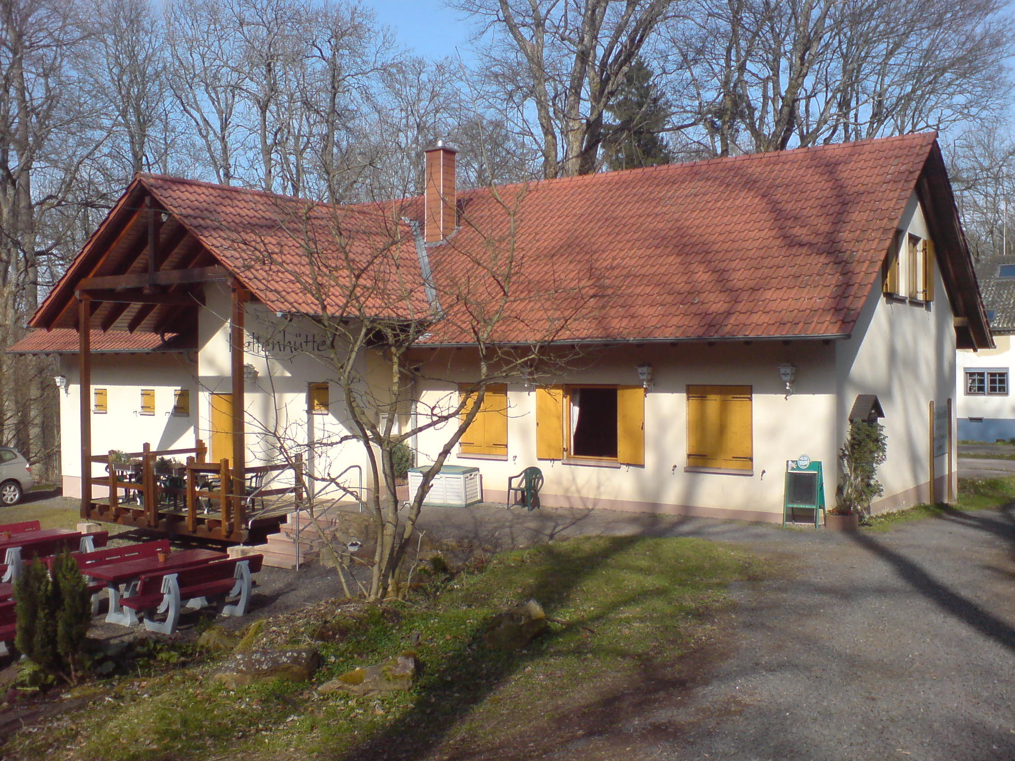 A one story alpine style building with a red tiled roof and light colored walls surrounded by bare trees in early spring.