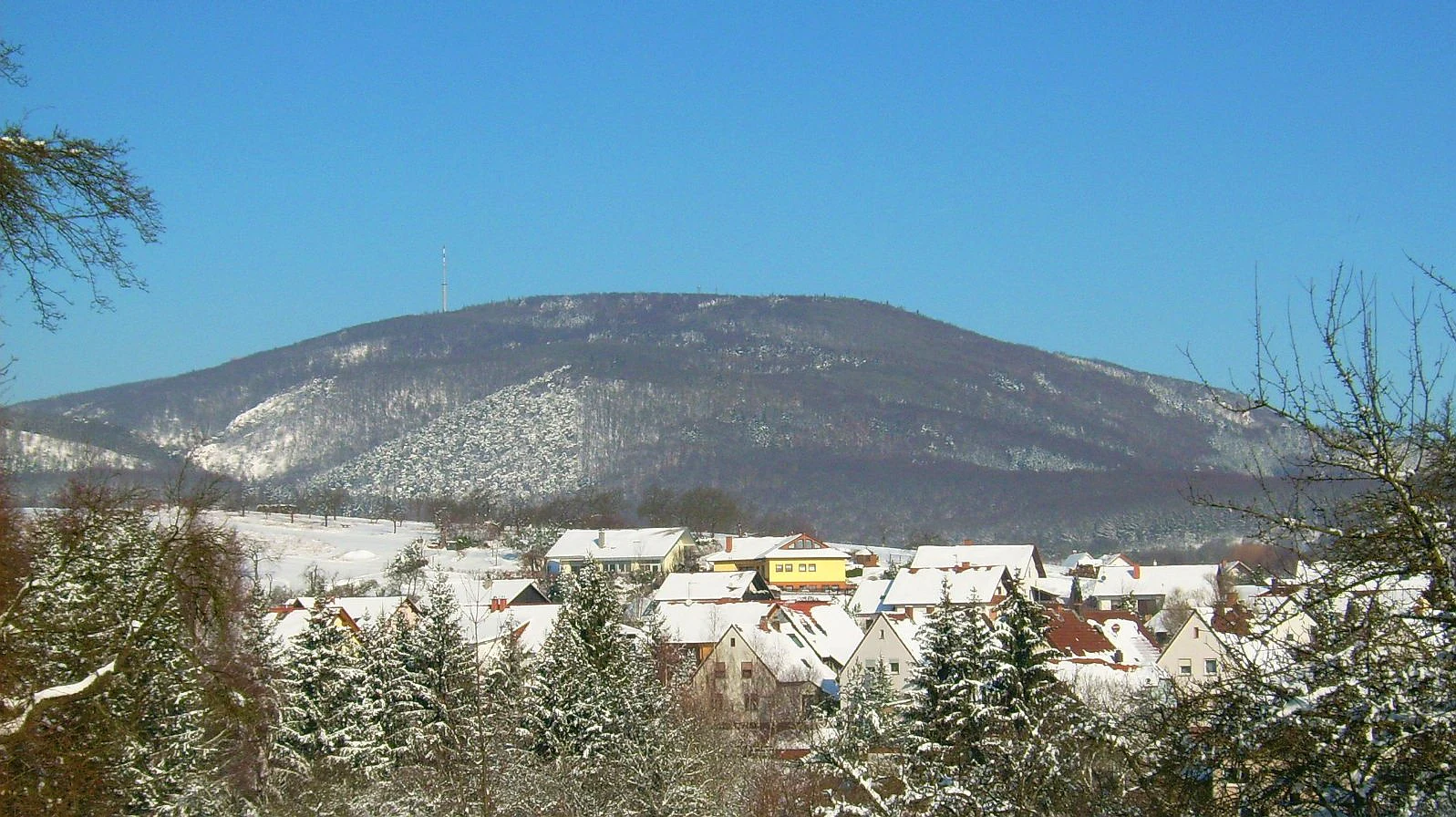 A wide view of a large, snow-covered mountain massif rising behind a small village, under a clear blue sky in winter.