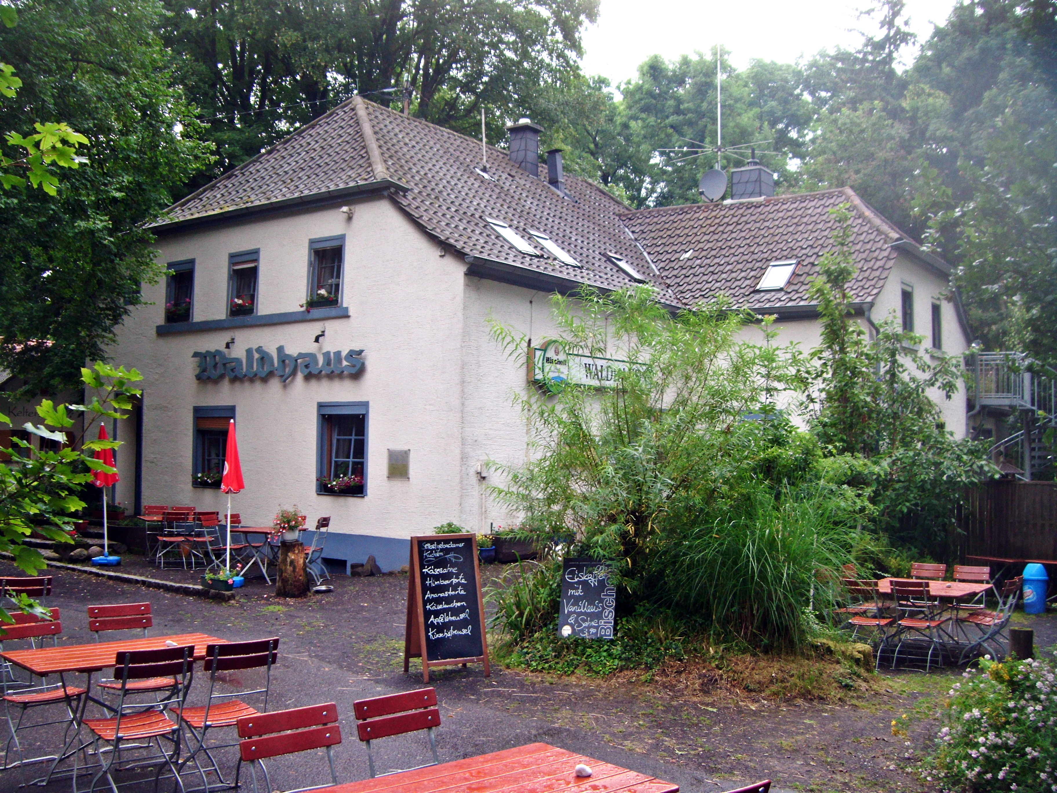 A two-story building named Waldhaus with outdoor seating, red tables, and chairs in a forest setting on Donnersberg mountain.