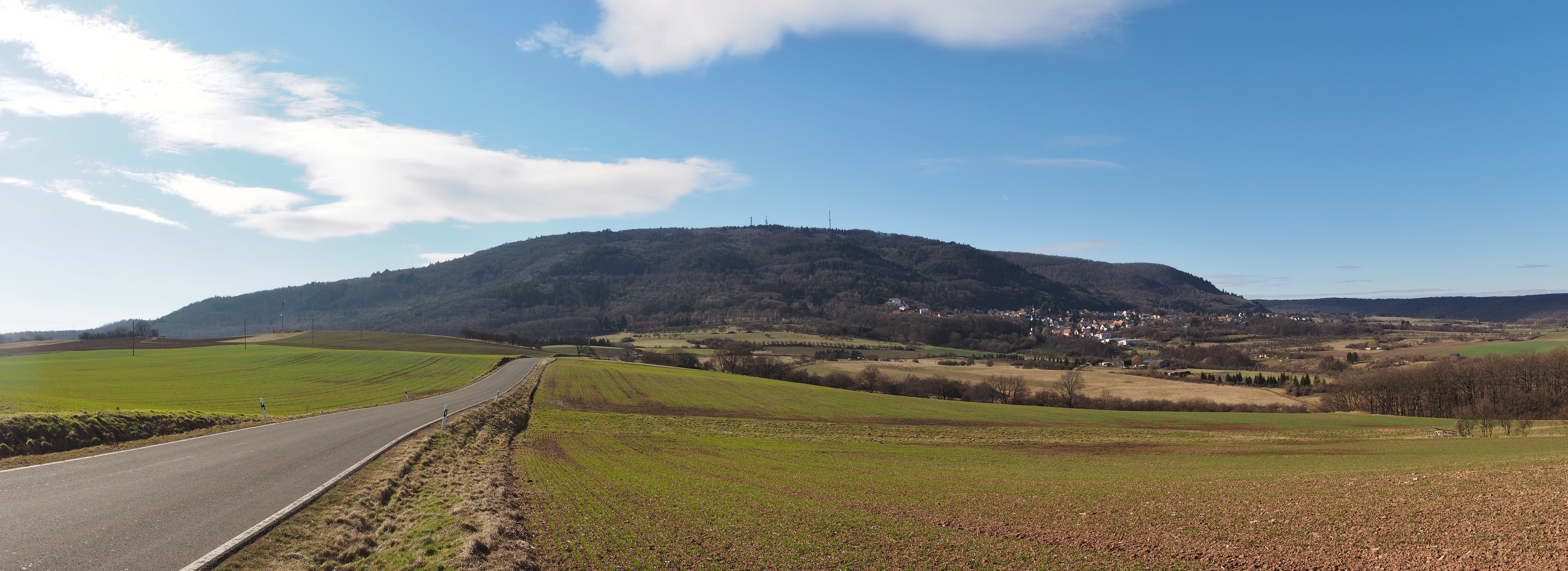 A wide, scenic view of the Donnersberg mountain range from the east, featuring a paved road in the foreground and rolling agricultural fields leading to the forested mountain slope.
