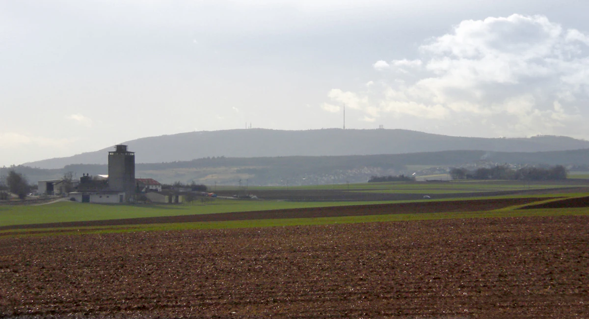 A wide landscape view of the Donnersberg mountain massif in Germany, seen from the east across open agricultural fields, with a farm building visible in the foreground left.