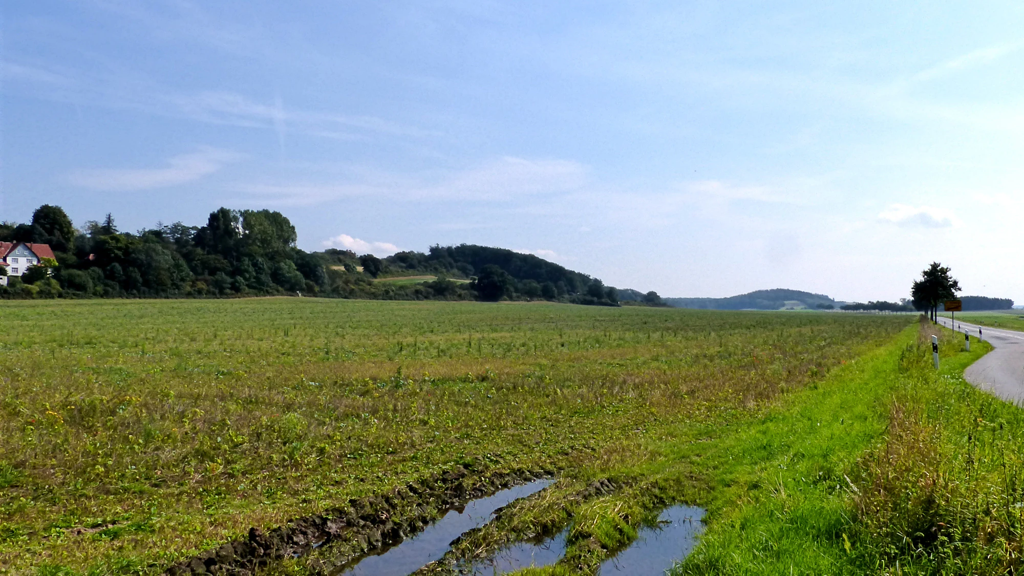 A wide, outdoor shot of a rural landscape with a large grassy field in the foreground, a paved road on the right, and a tree-covered ridge in the background under a blue sky.