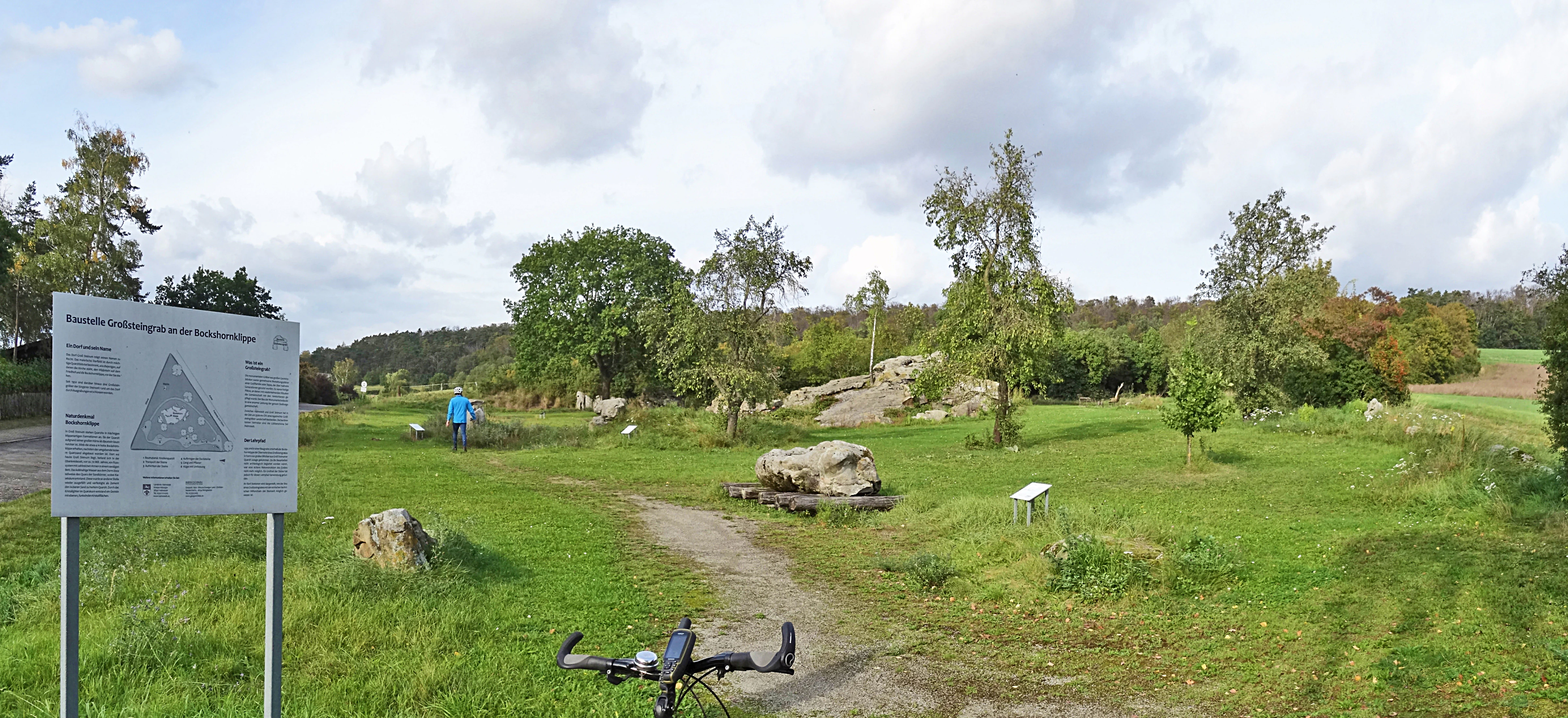 A white information sign stands in the foreground on the left, overlooking a grassy field with large boulders, scattered trees, and a dirt path leading toward the distance.