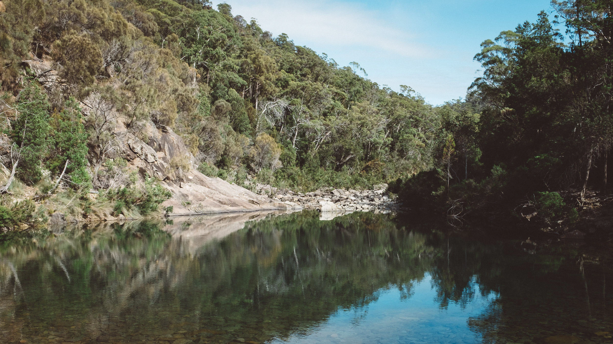Calm river reflecting rocky hills and dense forest under clear sky