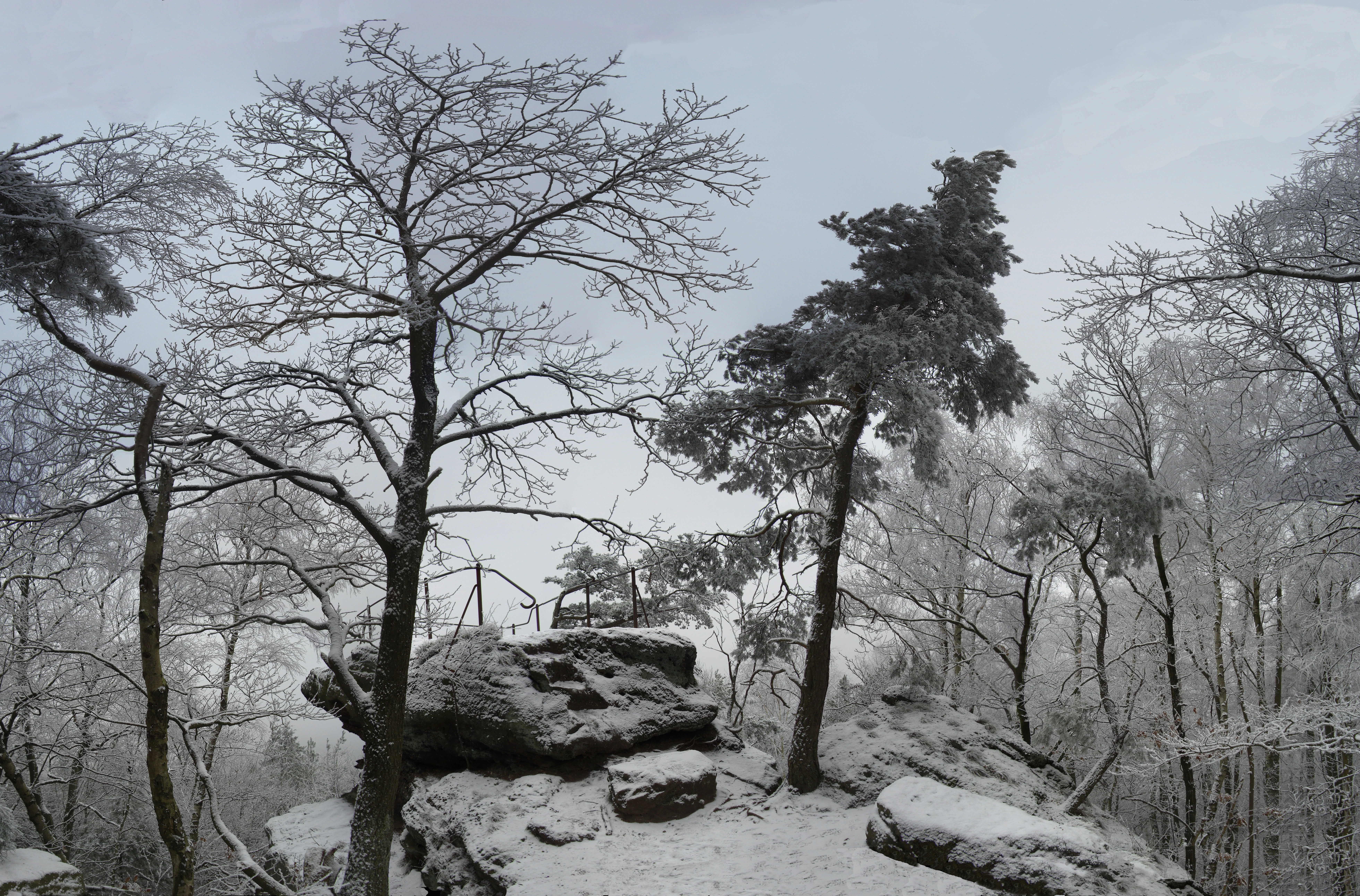 A winter scene at a rocky viewpoint featuring large sandstone boulders and bare trees dusted with snow under an overcast sky.