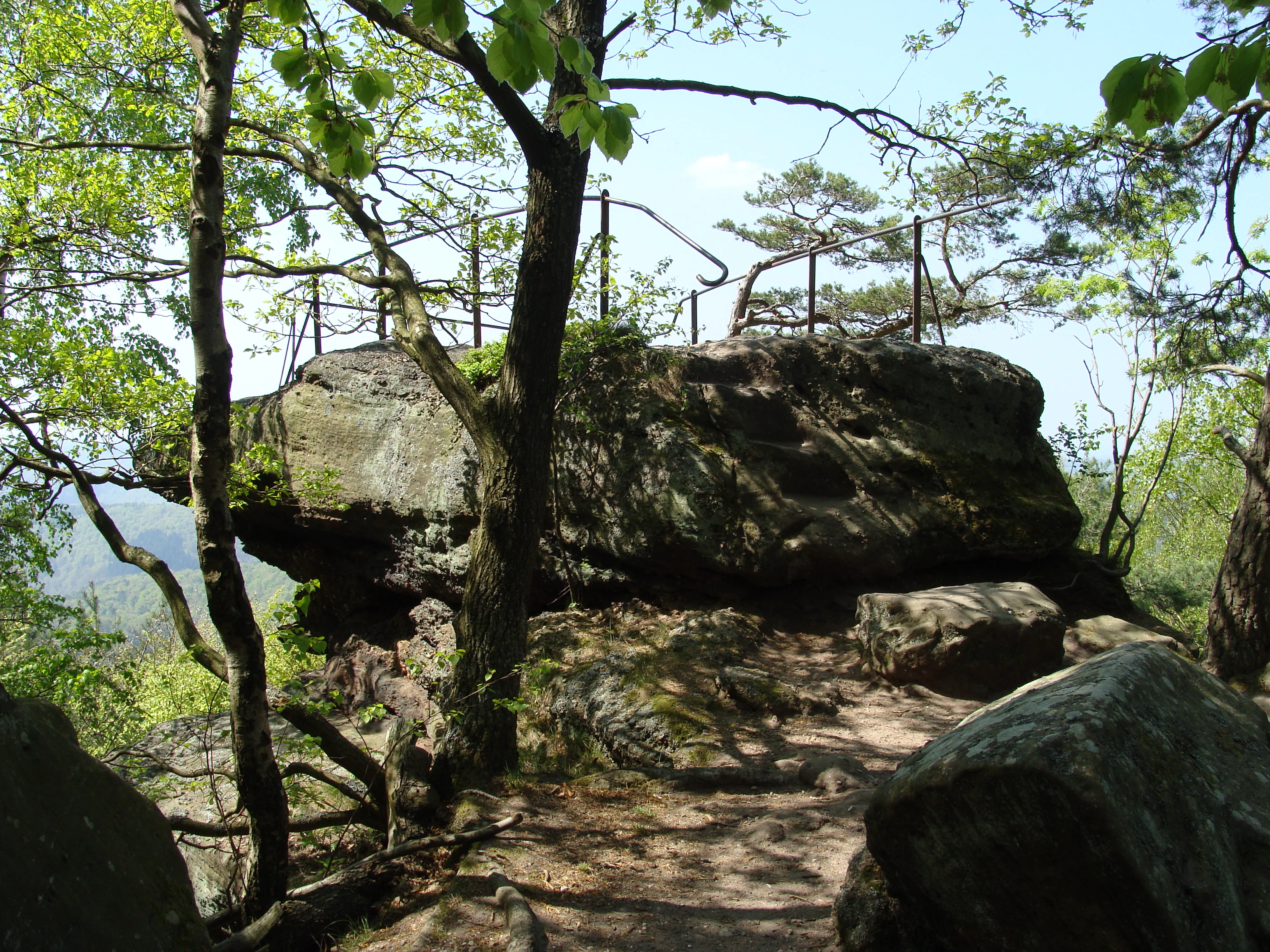 A large, weathered sandstone rock formation topped with a safety railing, surrounded by deciduous trees, serving as a viewpoint in the Palatinate Forest.