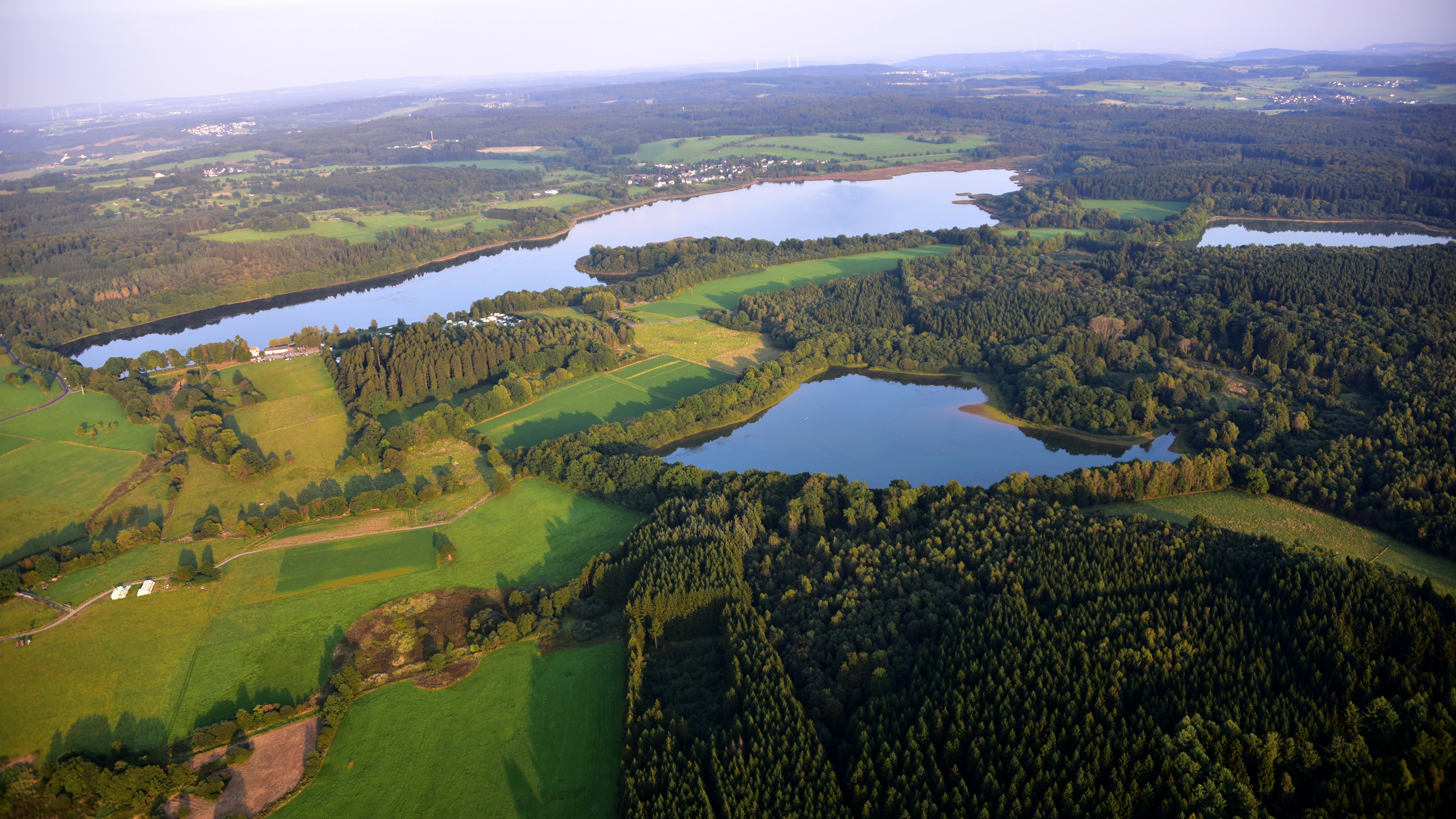 Perspectiva aérea mirando hacia abajo a un paisaje de lagos interconectados, densos bosques y parcelas de verdes campos de cultivo en la Alemania rural.