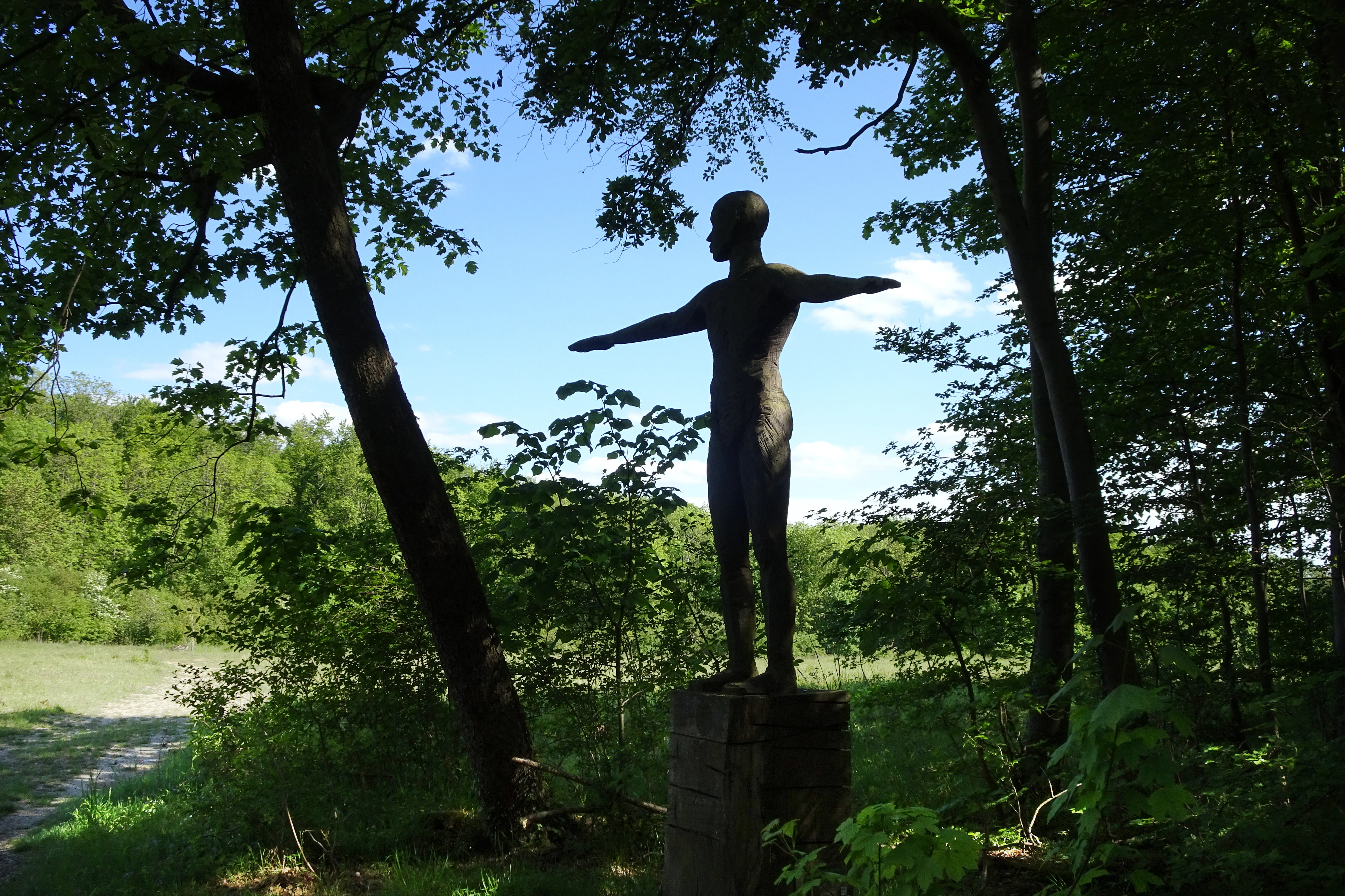 A dark human-shaped sculpture standing with arms outstretched on a pedestal in a sunlit forest clearing.