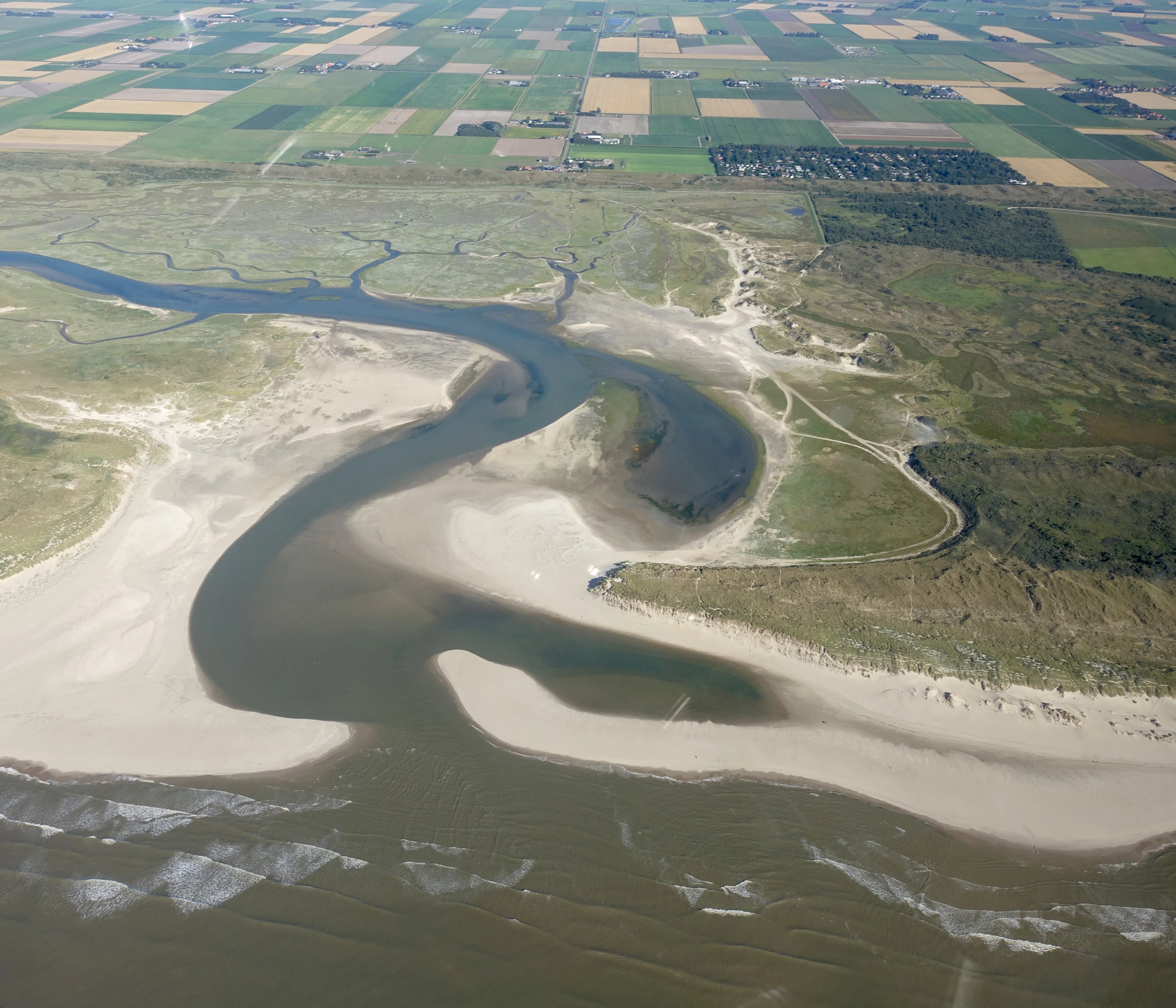Aerial view of a winding waterway surrounded by sandy dunes, coastal vegetation, and agricultural fields