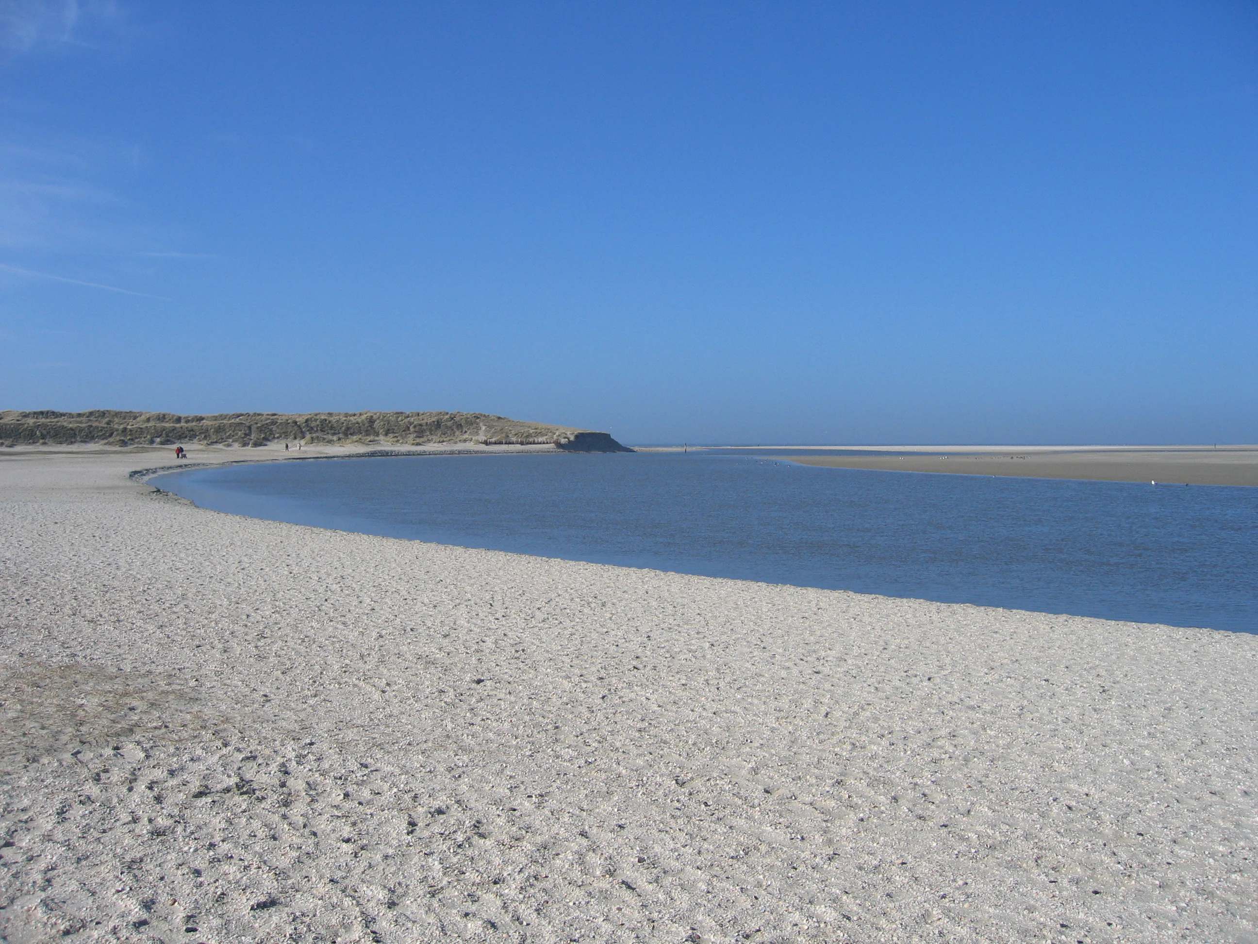 Wide sandy beach with calm blue water inlet under clear blue sky, with low dunes in background