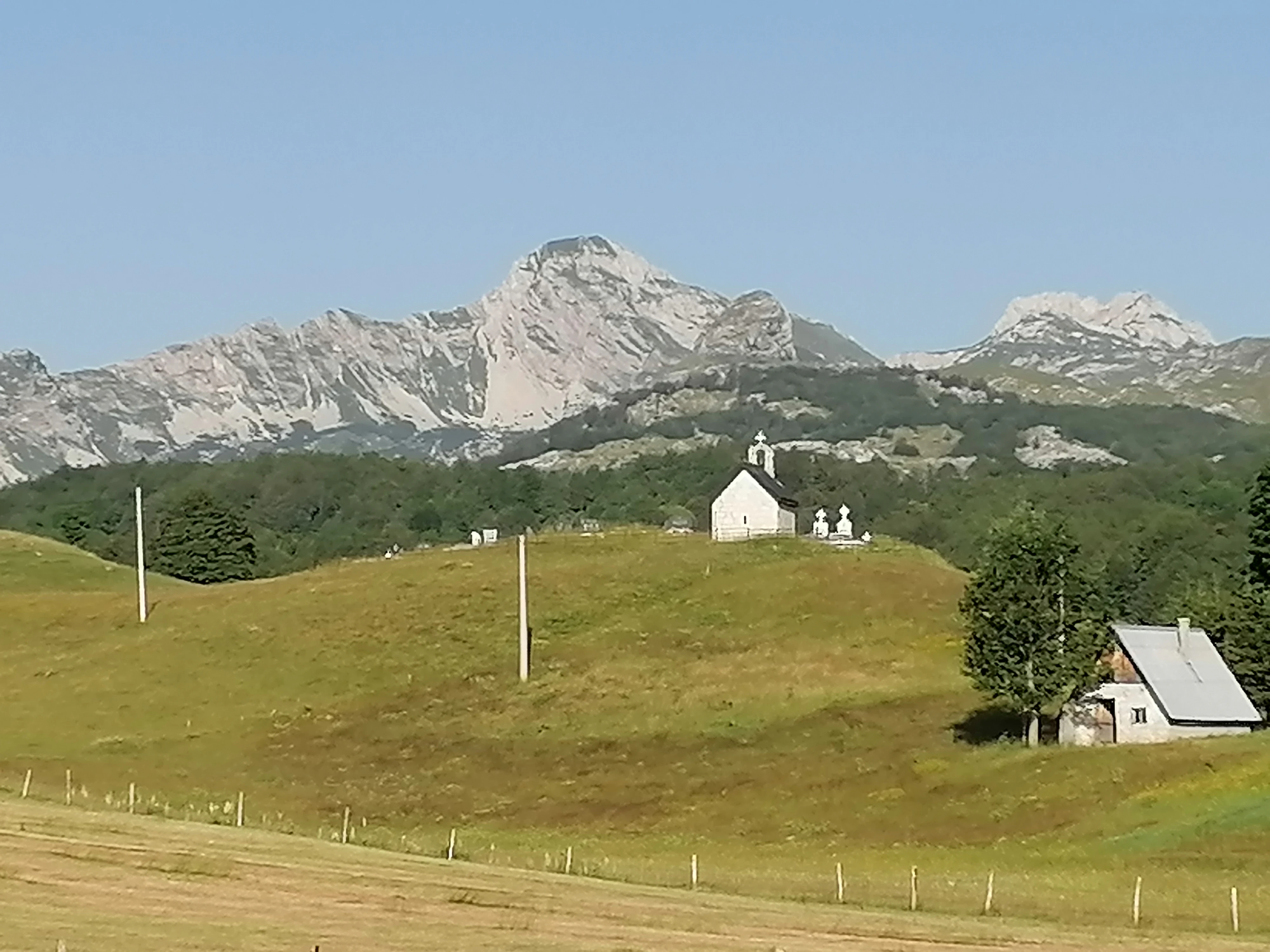 A small white chapel with a bell tower stands on a rolling green hillside, with a small house to the right and jagged, light-colored mountain peaks in the background under a clear sky.