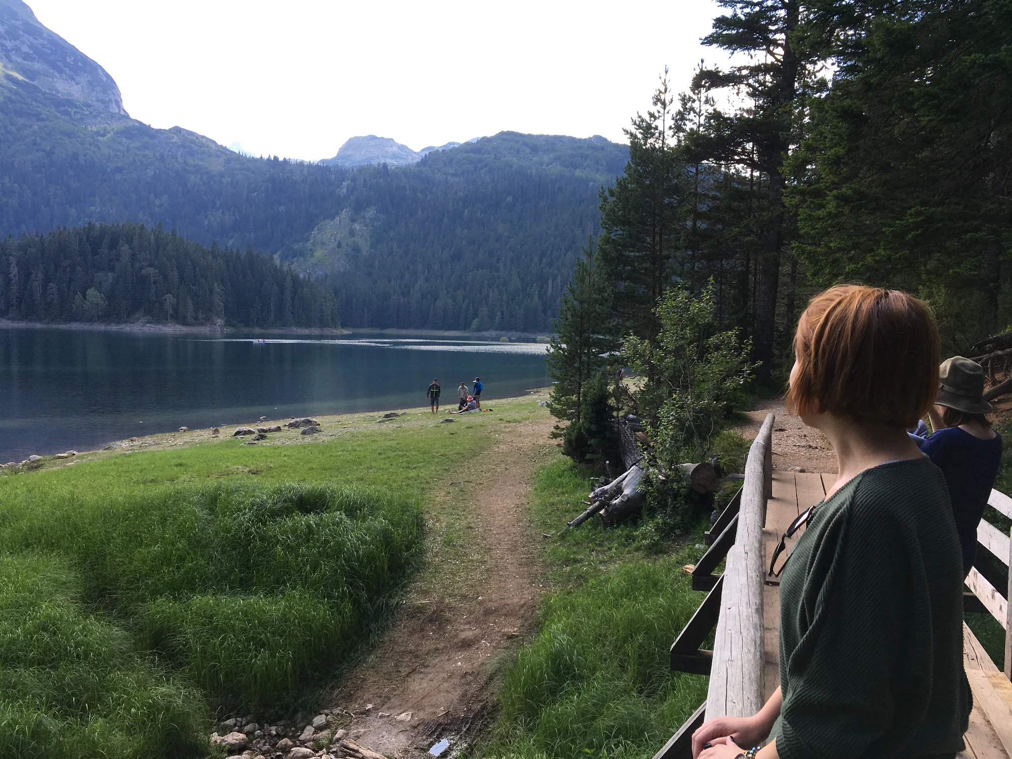 A woman with short hair stands on a wooden boardwalk, looking out over a calm alpine lake with forested mountains in the background under a soft sky.