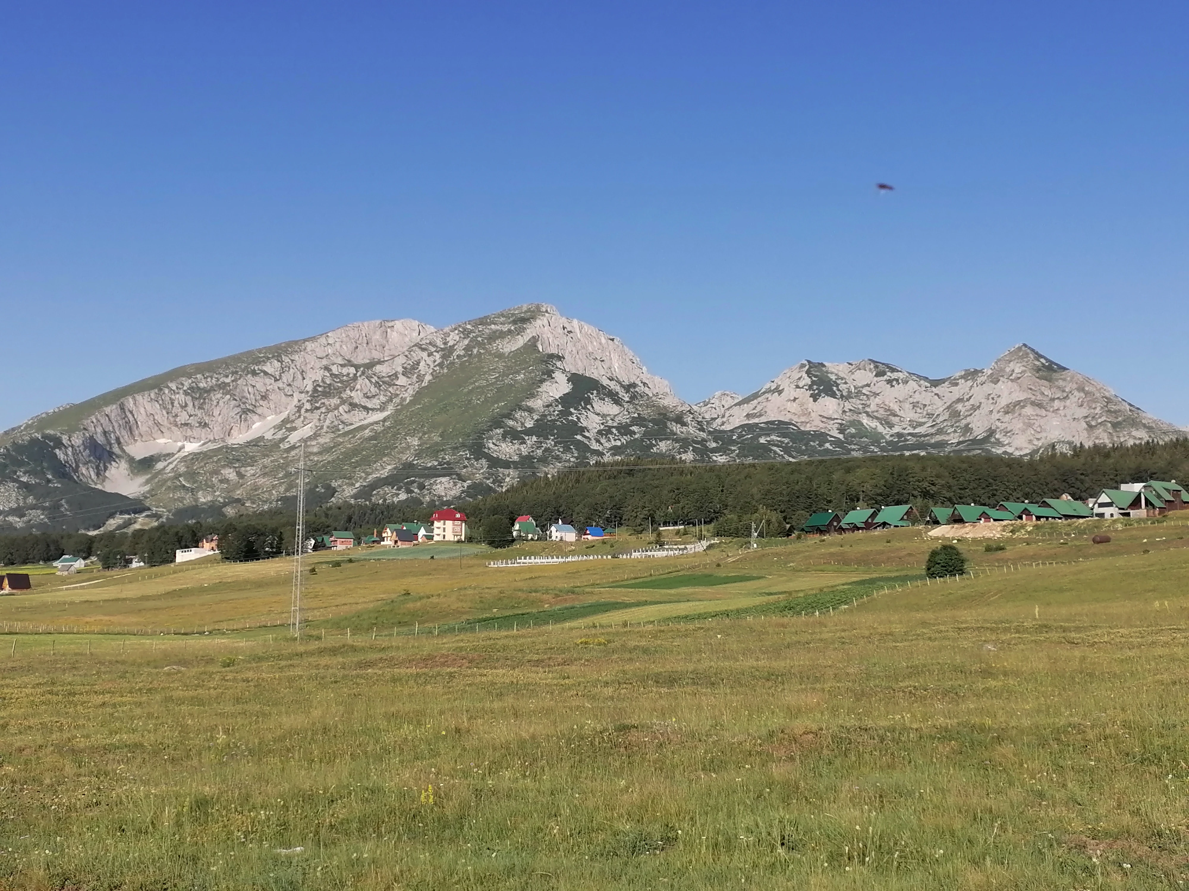 A wide landscape view featuring jagged rocky mountain peaks under a clear blue sky, with a green grassy field and a small cluster of buildings in the midground.