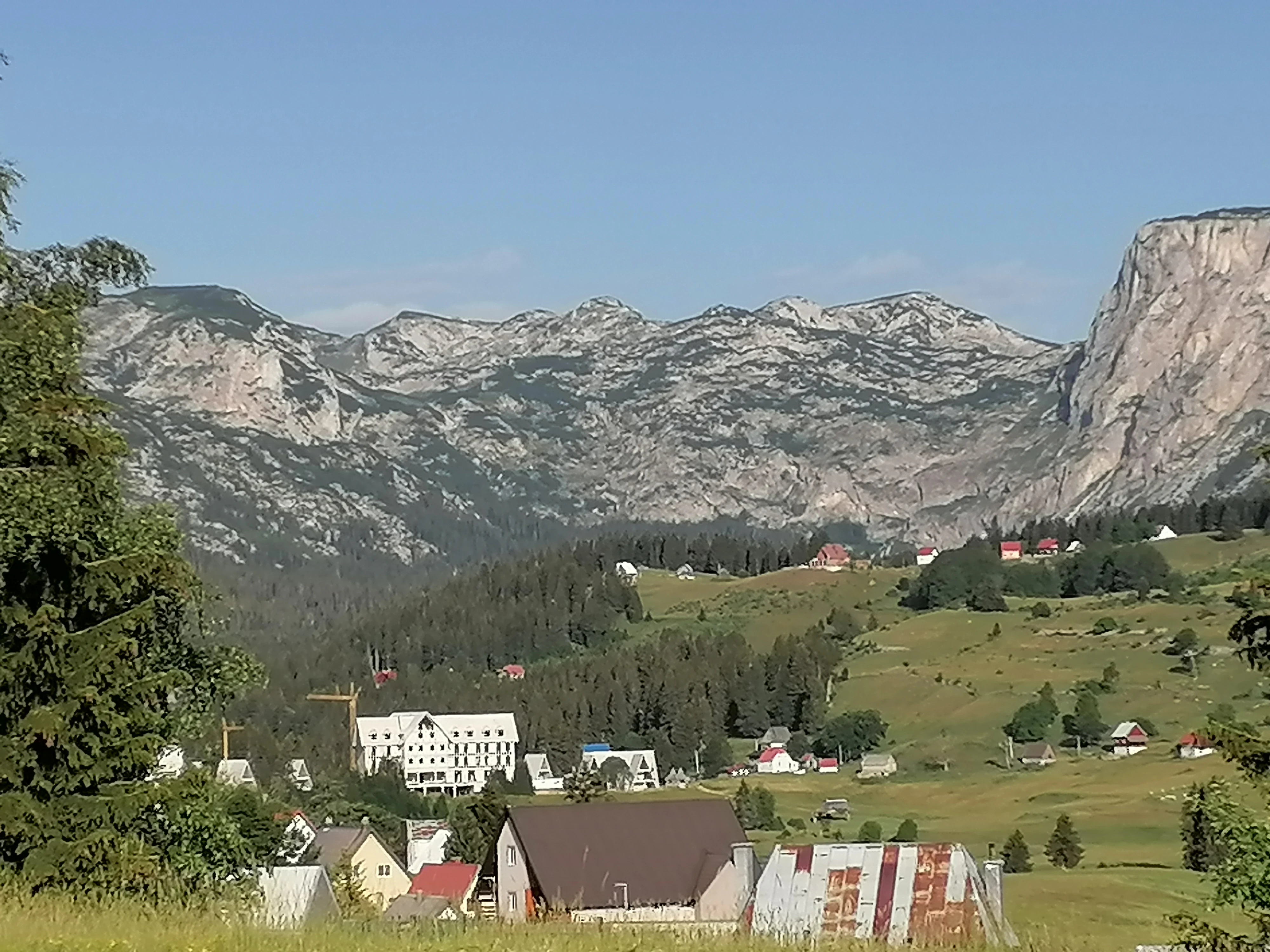 Mountainous landscape with rocky peaks, forested hills, and a village with scattered buildings under a clear blue sky