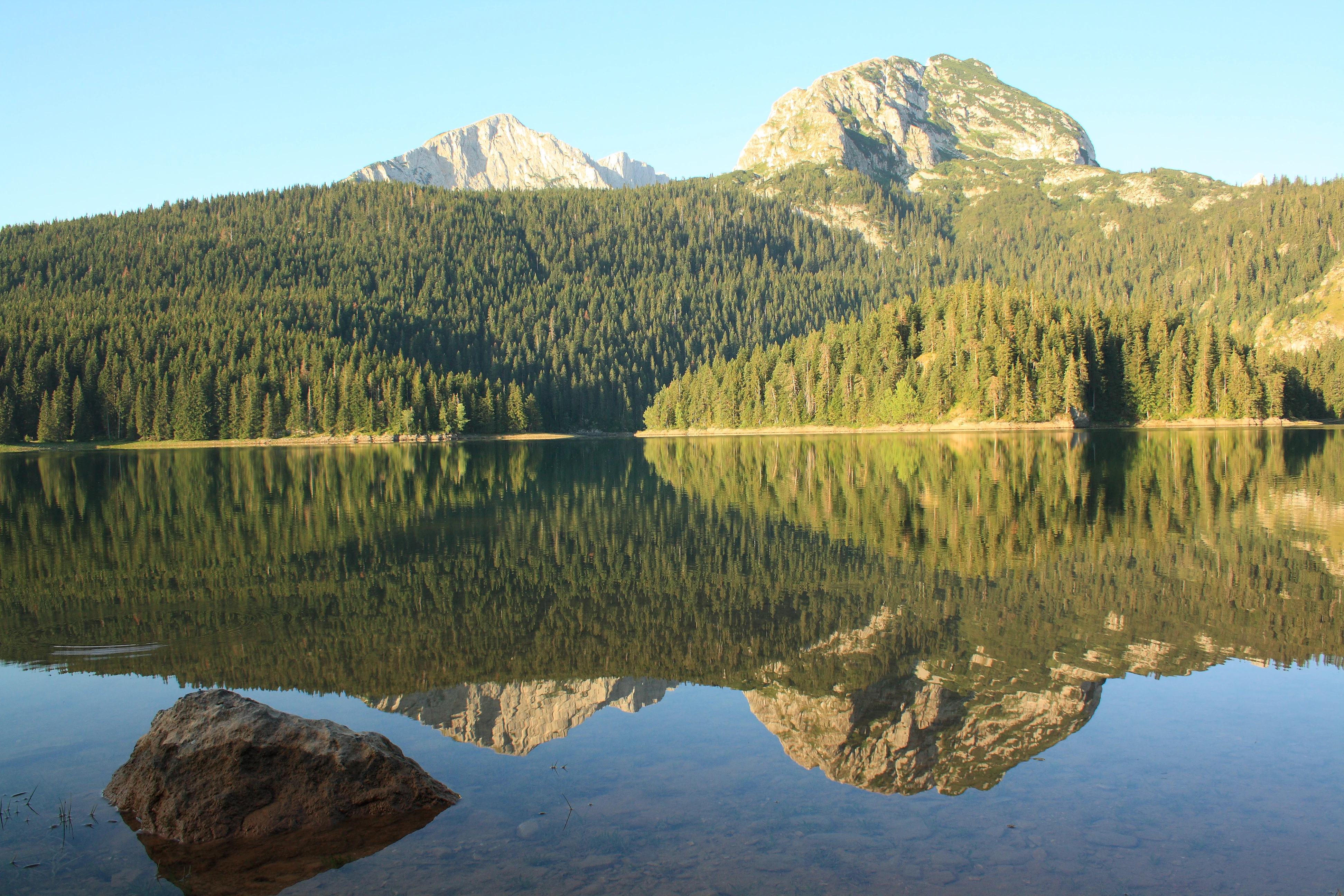 A calm lake reflecting mountains and forests with a large rock in the foreground