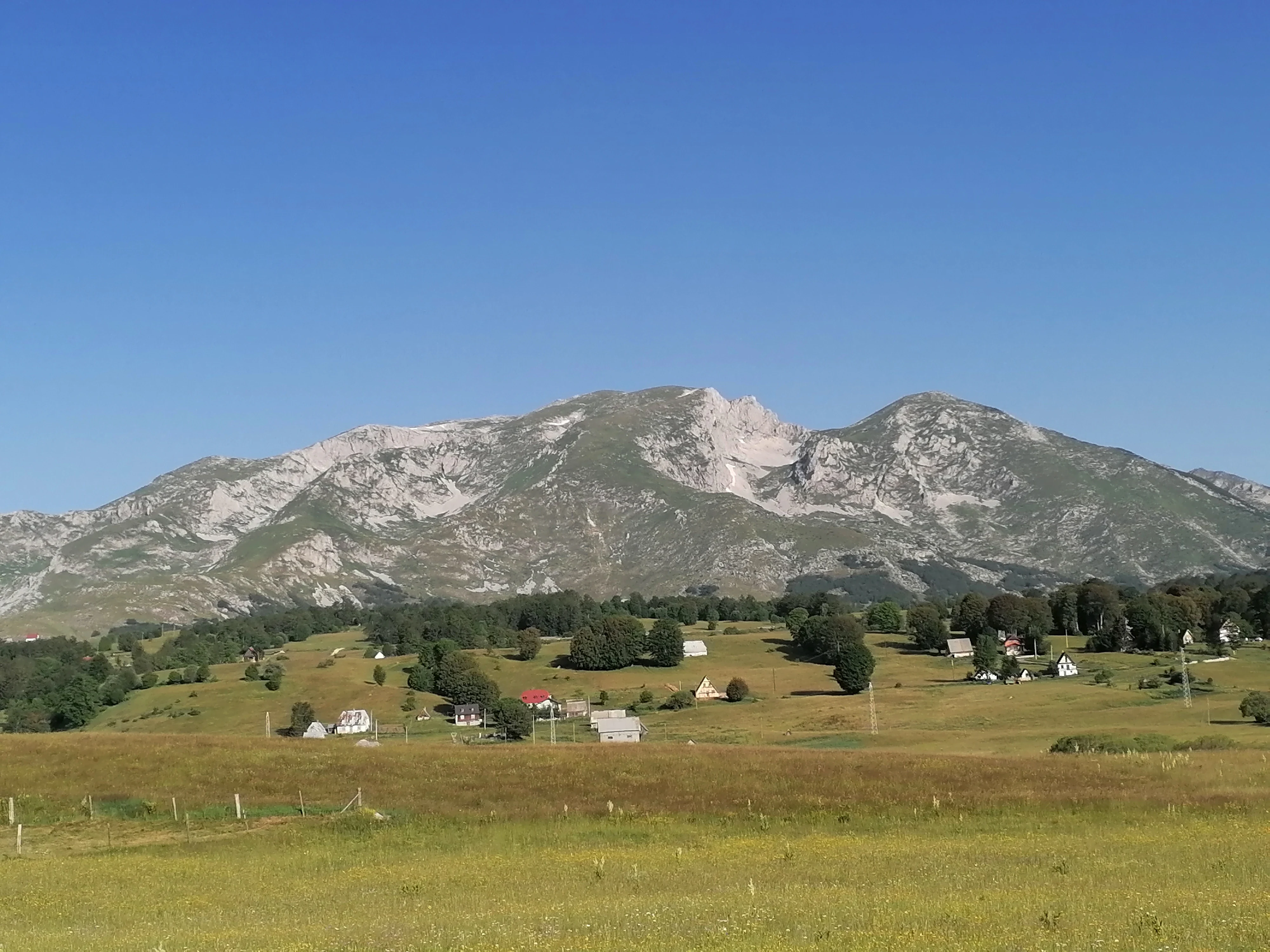 Wide view of Durmitor National Park showing a mountain, grassy field, houses, and trees under clear blue sky