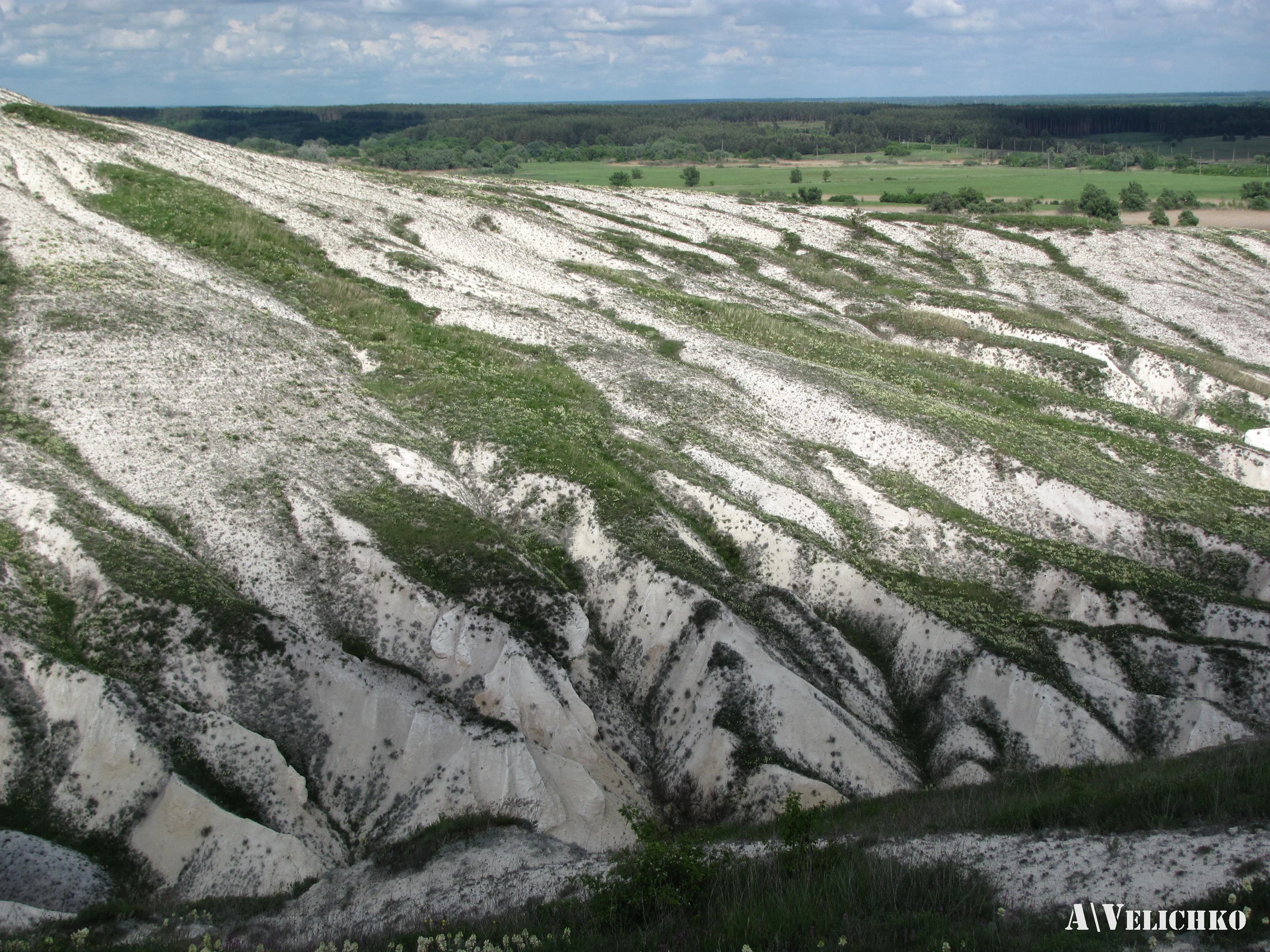 A wide landscape view of rocky hills with white and gray rock layers interspersed with green vegetation under a partly cloudy sky.