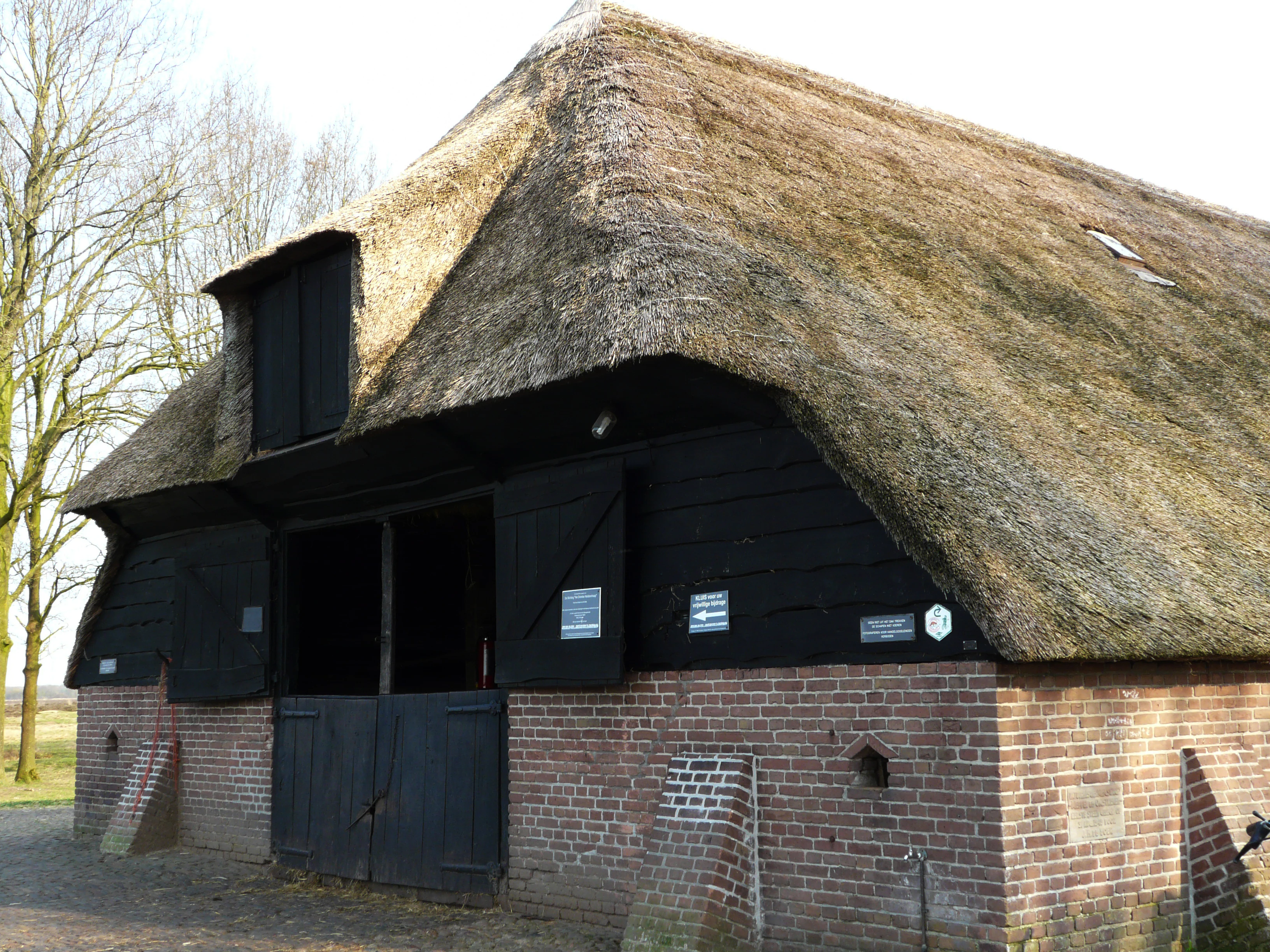 Traditional brick building with thatched roof and black wooden doors, featuring informational plaques on the exterior