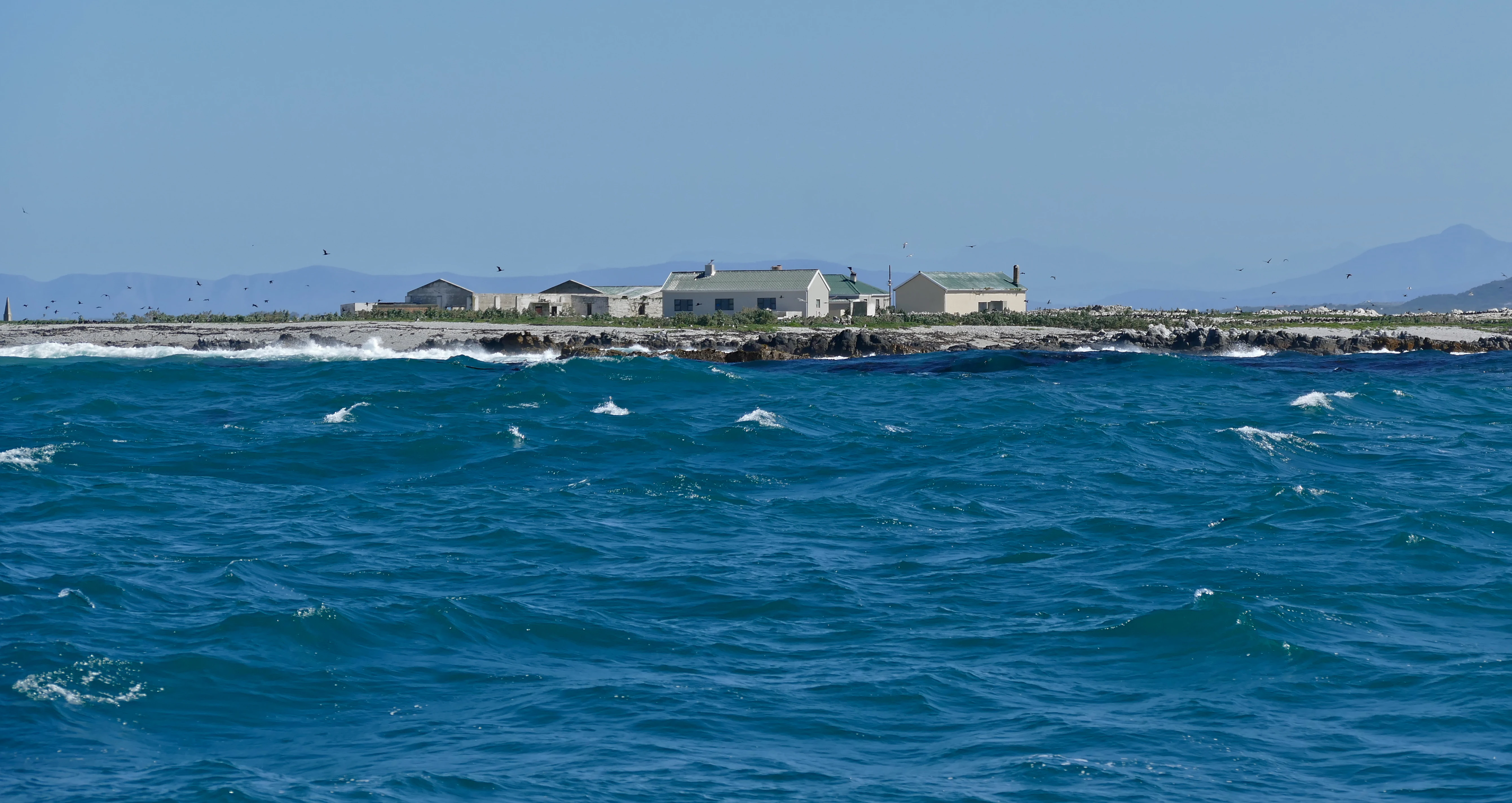 A low-lying island surrounded by blue sea, with small light-colored buildings situated on the rocky shore.