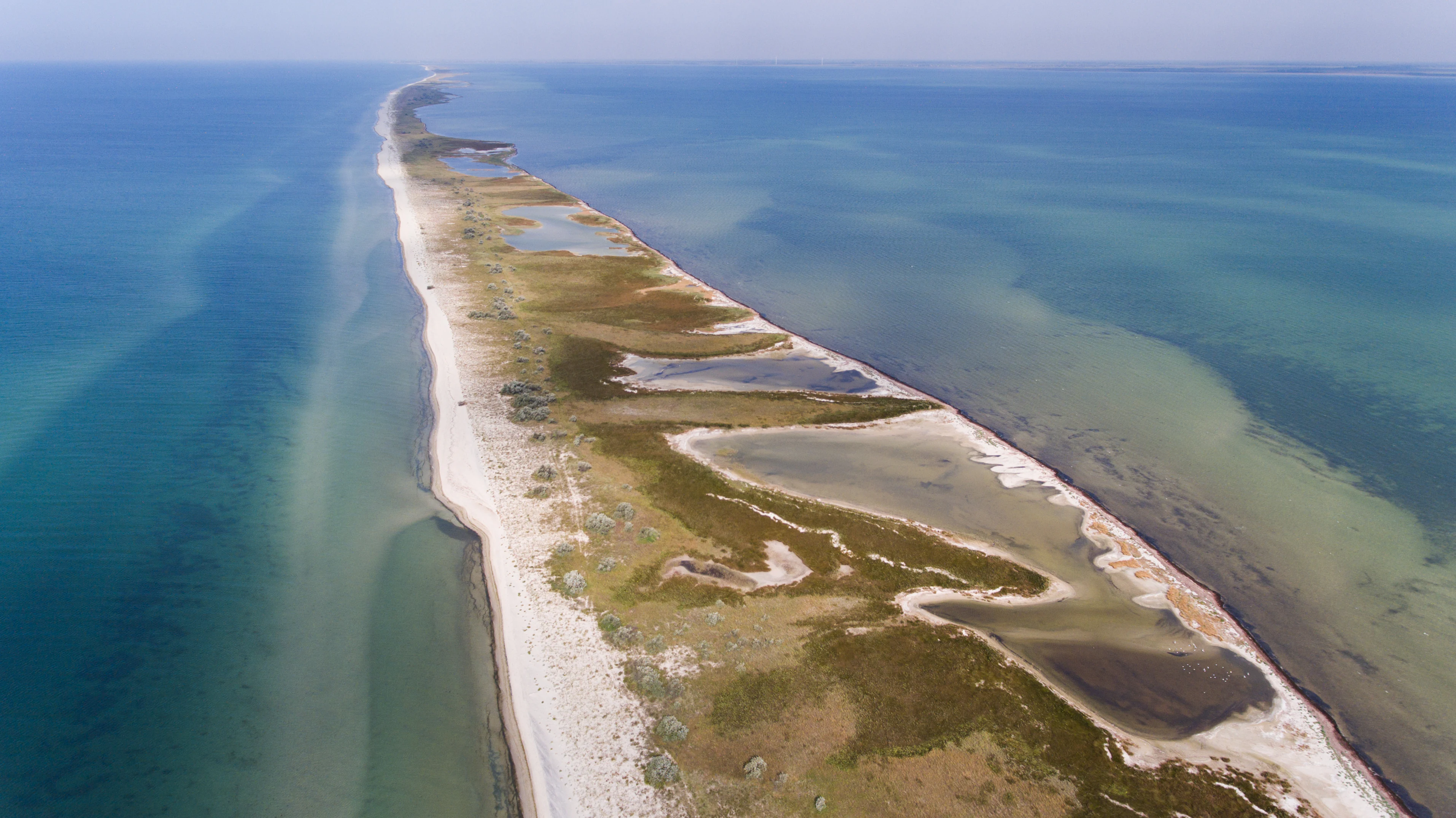 Aerial view of a narrow coastal spit with sandy and grassy terrain surrounded by shallow blue-green waters