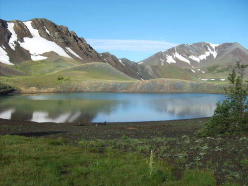 A calm, circular alpine lake reflecting snow-dusted mountain slopes under a clear blue sky.