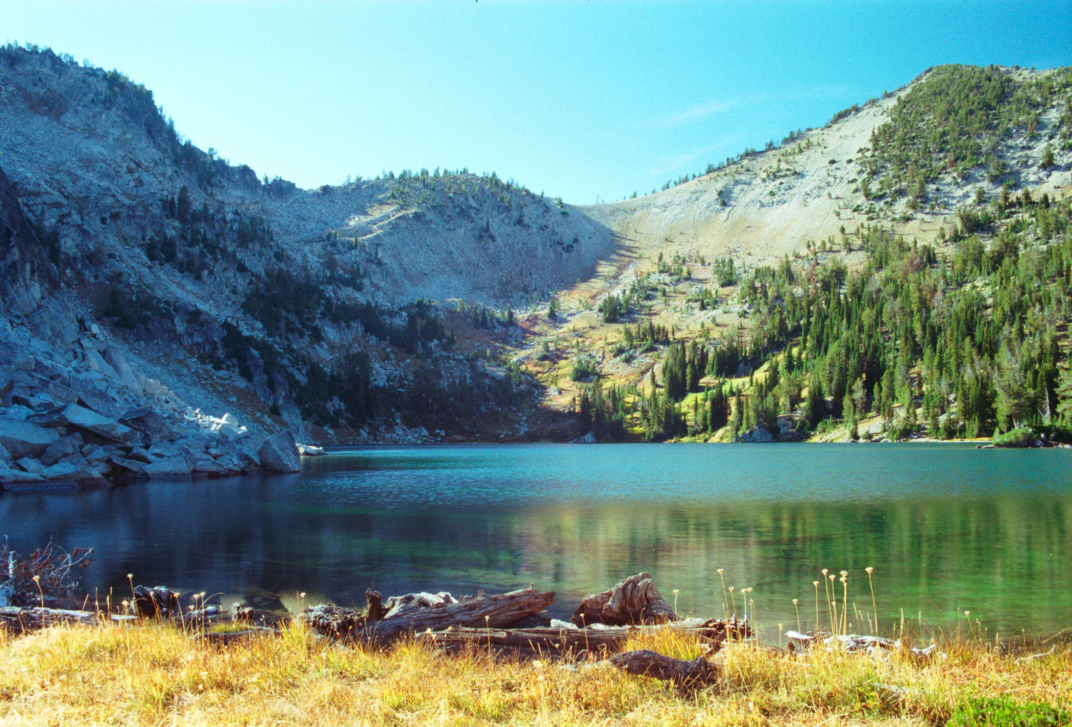 A clear, still alpine lake, Maxwell Lake, sits in a glacial basin surrounded by rocky, scree-covered peaks and a coniferous forest under a clear blue sky.