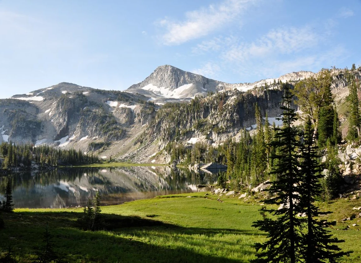 A clear, calm mountain lake reflects jagged, snow-dusted granite peaks in the background, with a vibrant green meadow and evergreen trees in the foreground.