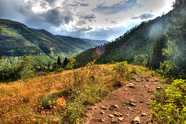 A dirt hiking trail leads through golden meadows and green mountain slopes under a cloudy sky.