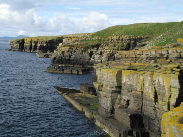 Rugged, layered sandstone cliffs along a coastline, with green grassy slopes above and dark blue ocean water below.