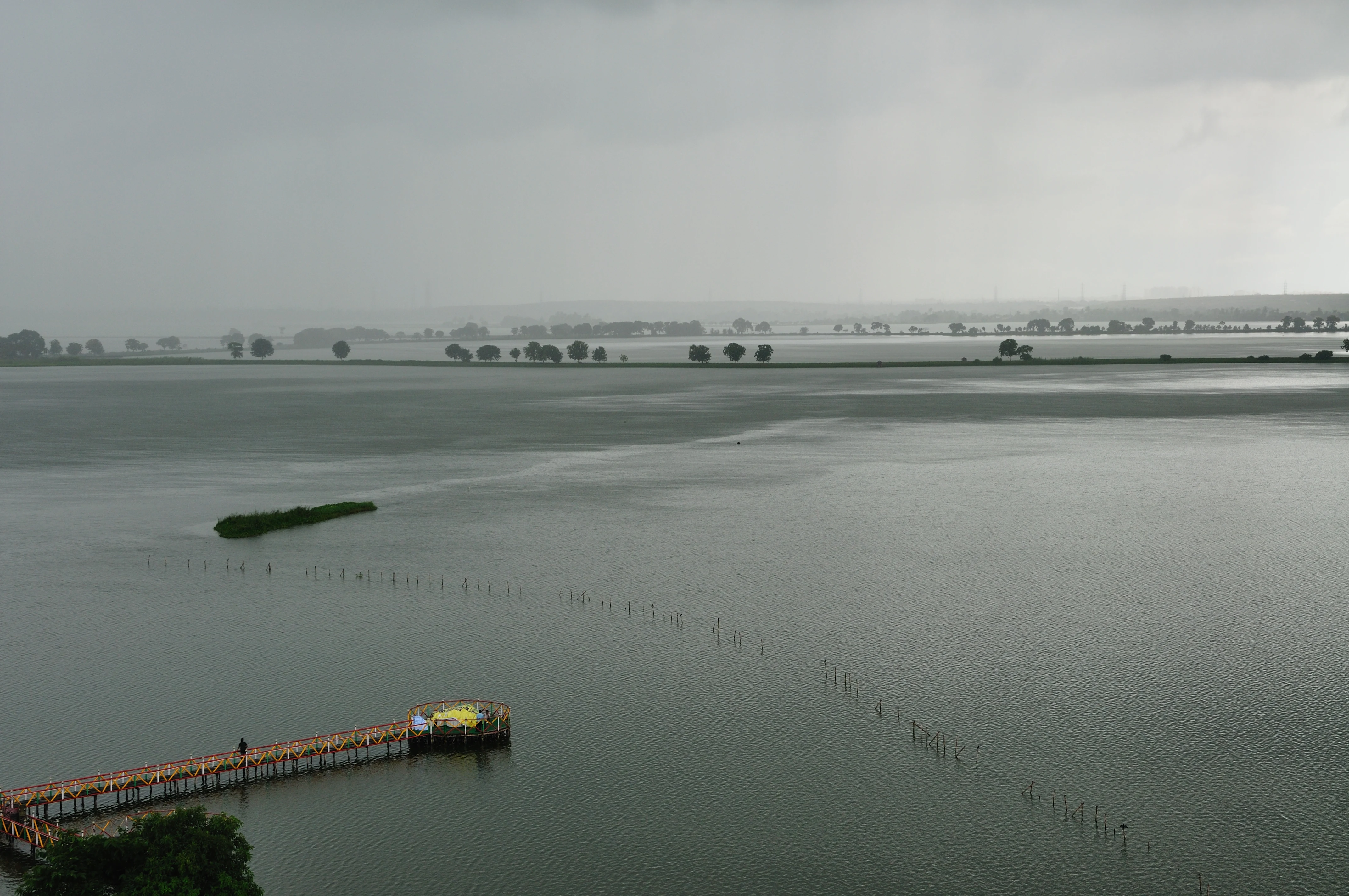 A wide, elevated view of a large, calm lake under a gray, cloudy sky. A long wooden pier extends into the water from the bottom left corner, and a series of thin, vertical posts are visible rising out of the water in a line stretching toward the horizon.