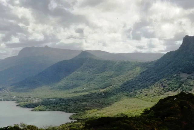 Vista panorámica de exuberantes laderas montañosas verdes que descienden hacia una bahía con cielos nublados.