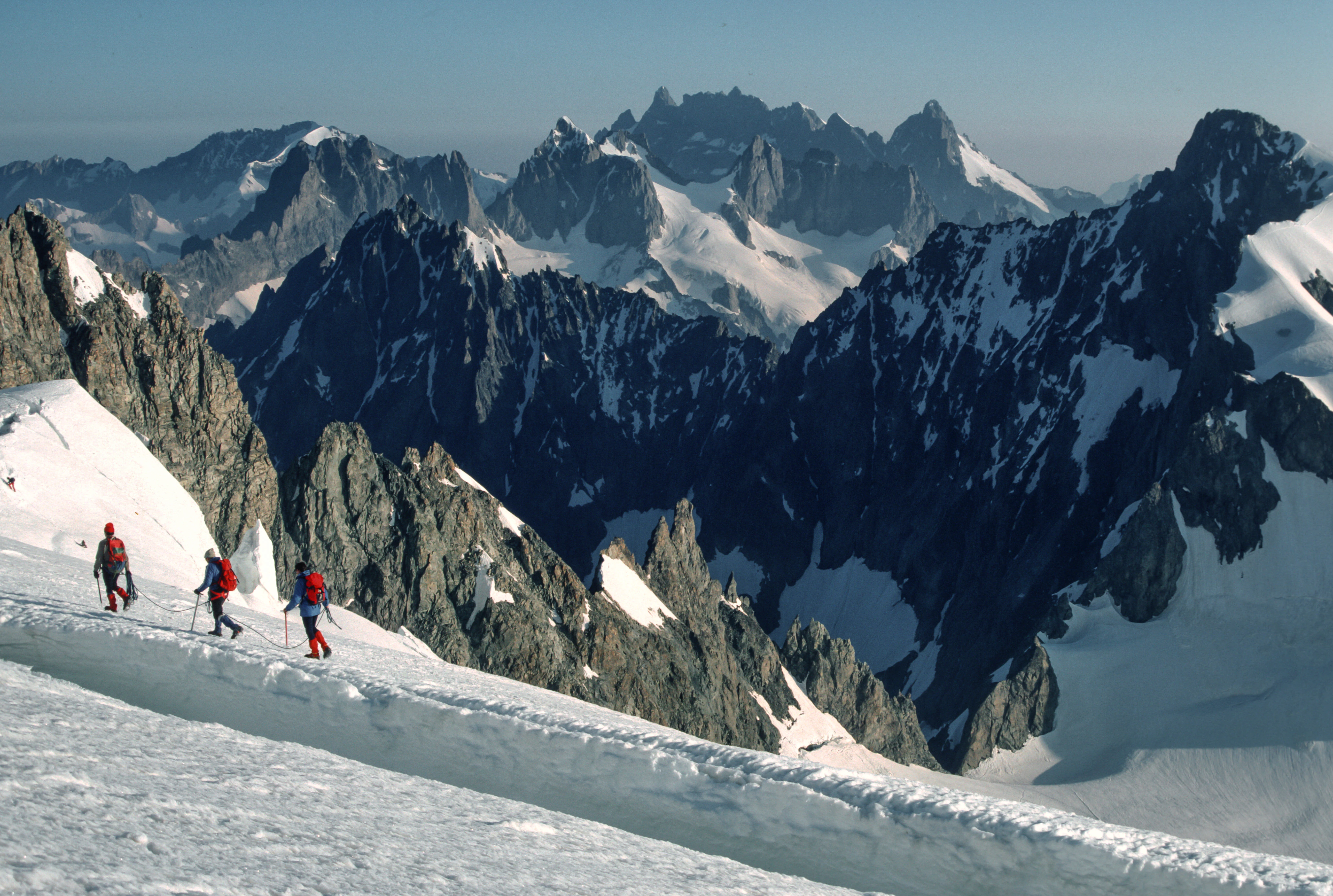 Three climbers in red gear ascending a snow-covered mountain slope with rugged rocky peaks and glaciers in the background