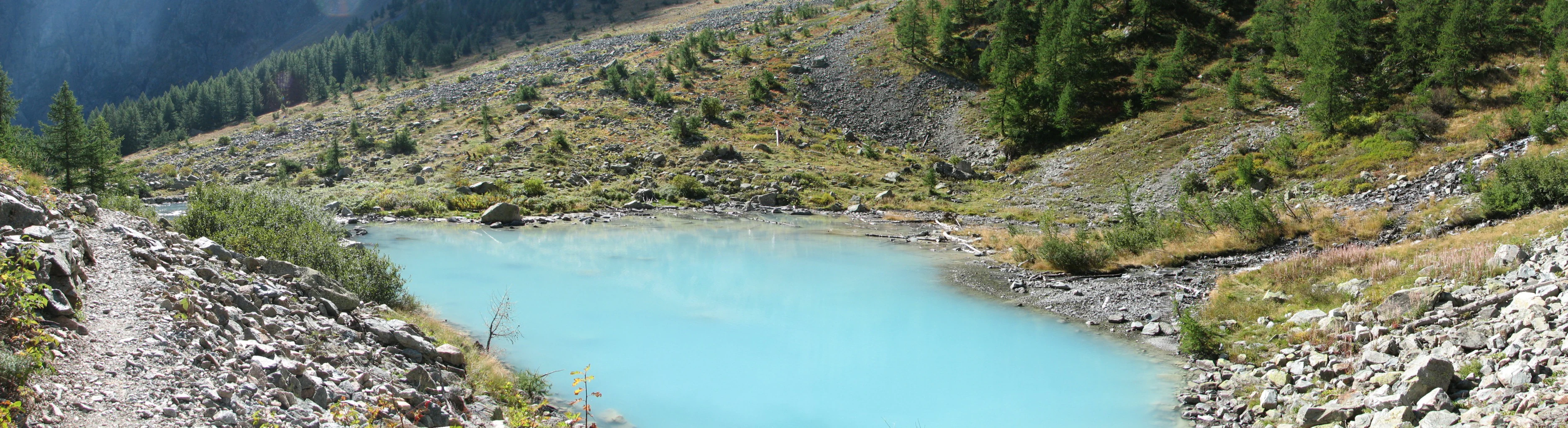 A turquoise lake surrounded by rocky terrain and a dirt trail, with forested hills and mountains in the background